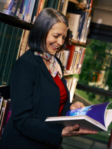 A woman in a dark blazer and colorful scarf smiles while holding an open book in a library. She has shoulder-length dark hair with some gray and appears to be enjoying the book, surrounded by shelves filled with various books in the background.