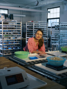 A woman in a pink shirt is sitting at a table in a laboratory filled with various equipment. Behind her are metal racks with numerous containers. She has her arms resting on the table, which has a green basket, glasses, and a weighing scale. The setting is well-lit, showcasing the organized workspace.