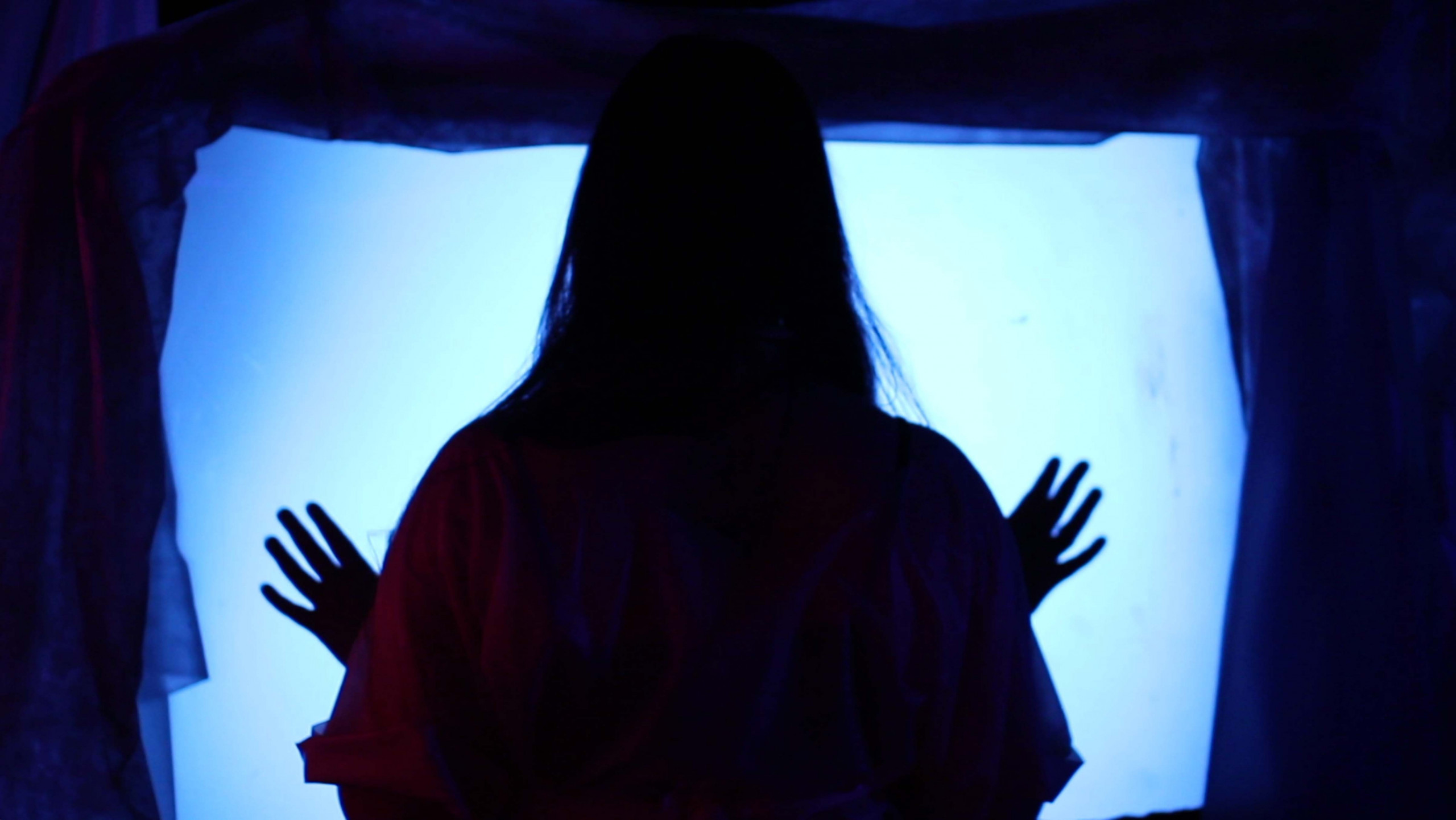 Silhouetted figure with long straight hair holds hands up to an illuminated blue-tinted screen