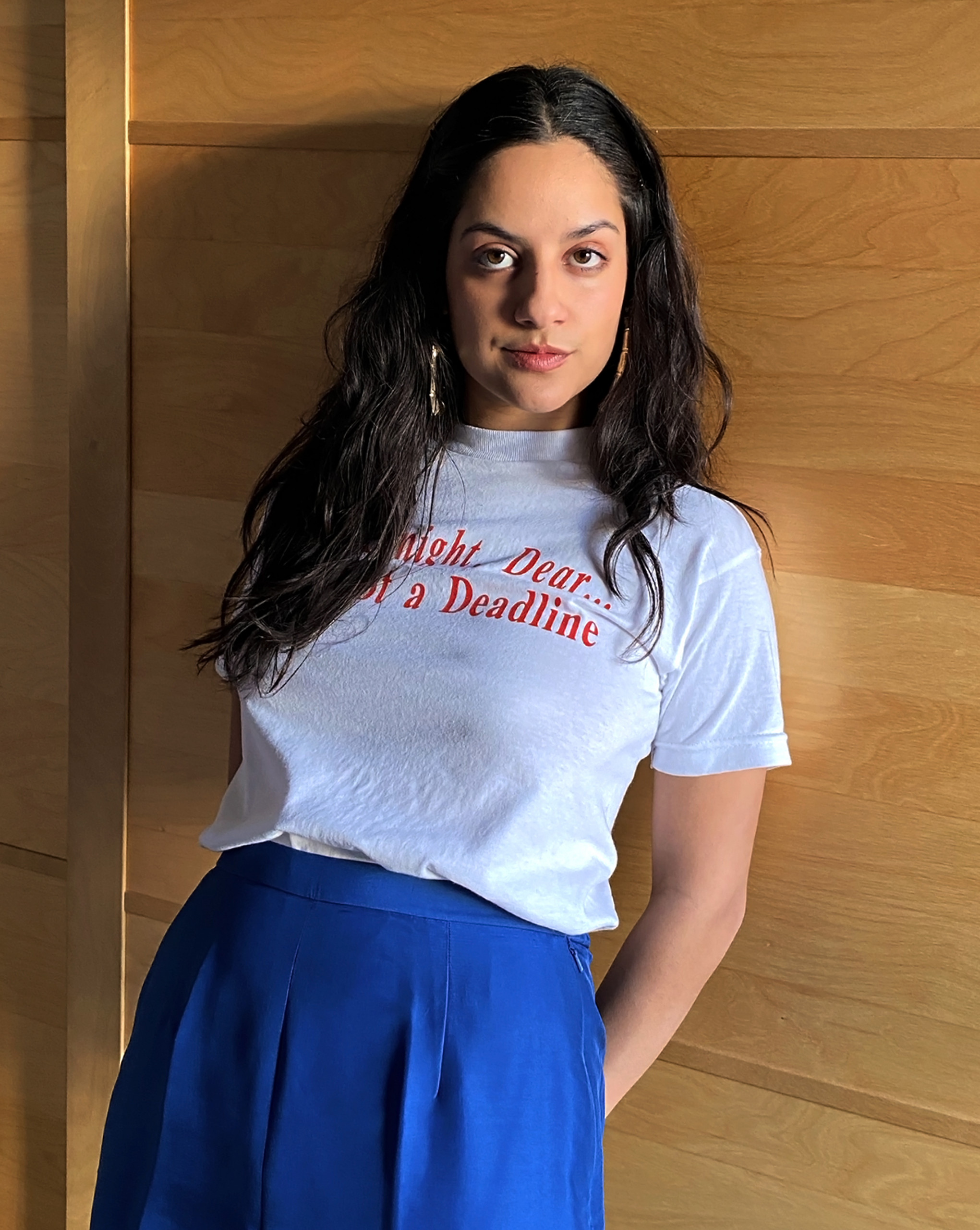 Headshot of Farah Kafei, with long dark brown hair, a white T-shirt with partially covered red lettering, and long gold earrings, leaning against a wood wall