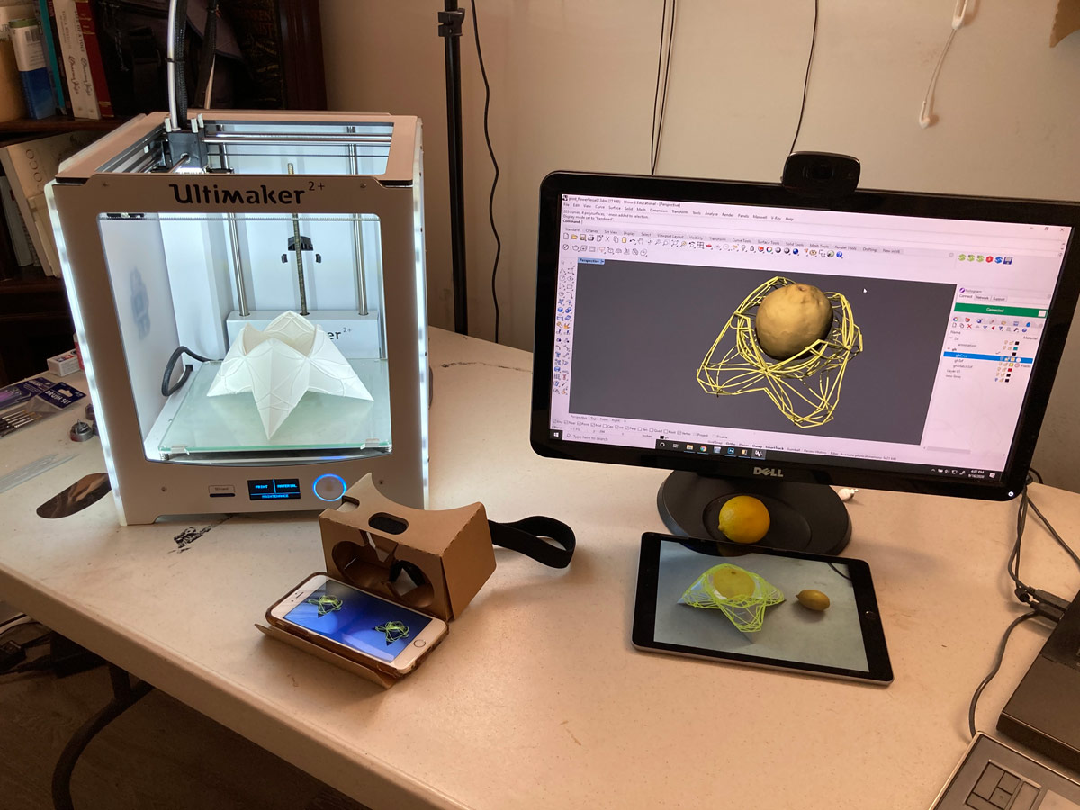 Desk with 3-D printer, augmented reality smartphone display, screens showing multiple views of a design object