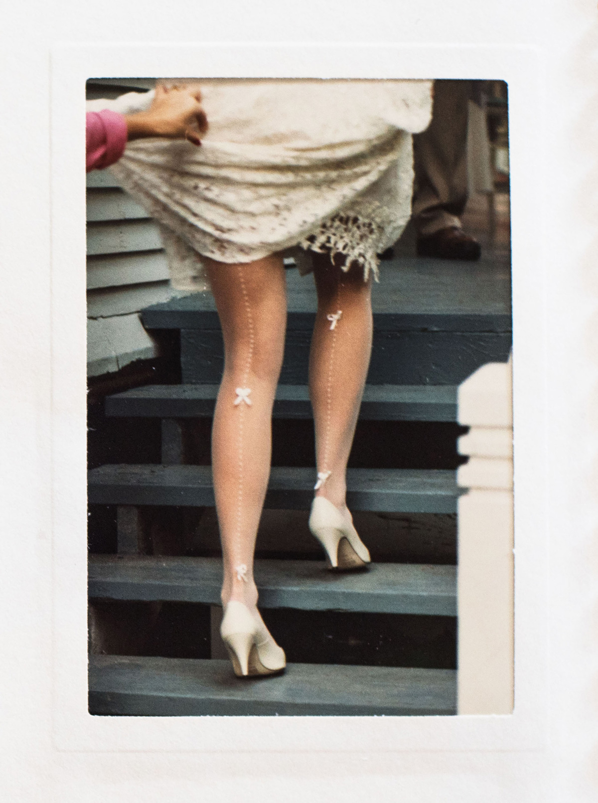 Picture of a woman in white heels, with her dress held up by another individual in pink while she walks up some steps