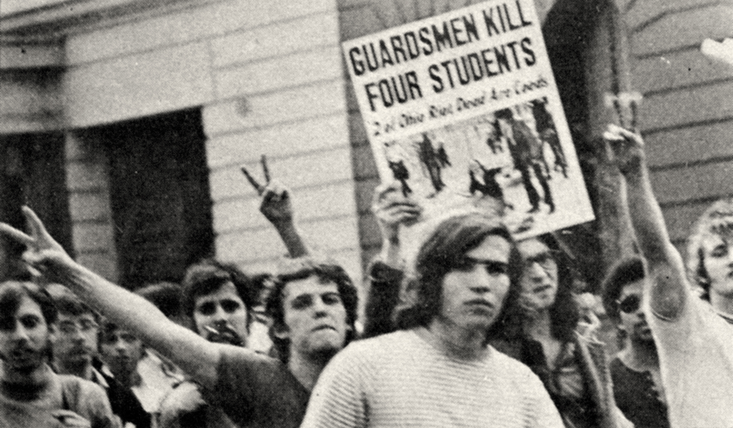 Students march holding up fingers in peace signs and carrying a paper reading Guardsmen Kill Four Students