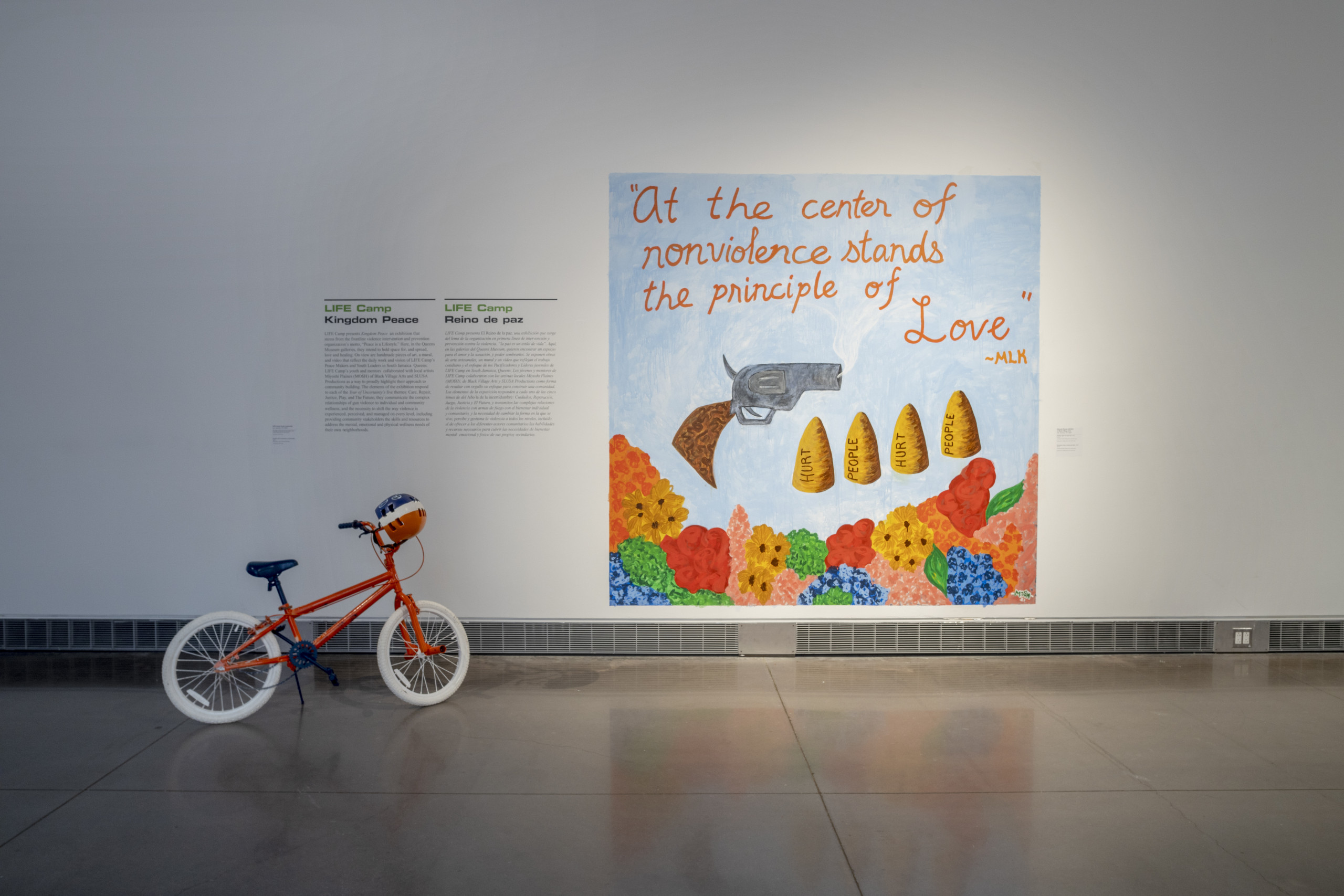 Mural with gun and oversize bullets floating above a field of colorful flowers, with wall text and a bicycle in the foreground