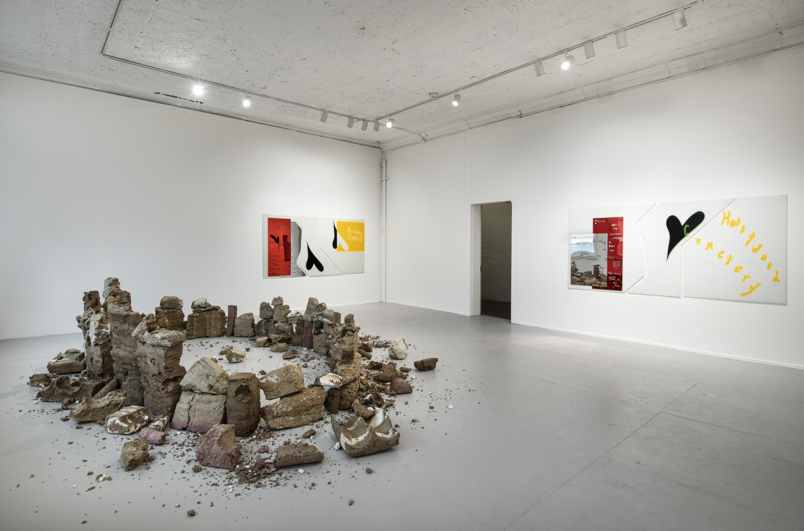 Gallery installation with fragmented sculpture foregrounding two white, red, mirrored, black, and yellow acrylic works on white walls