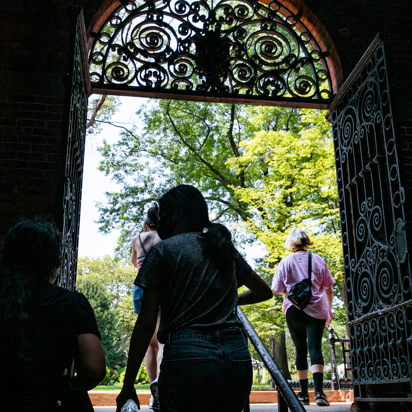 Students walk from shaded courtyard into sunlit campus under ornate metal gateway