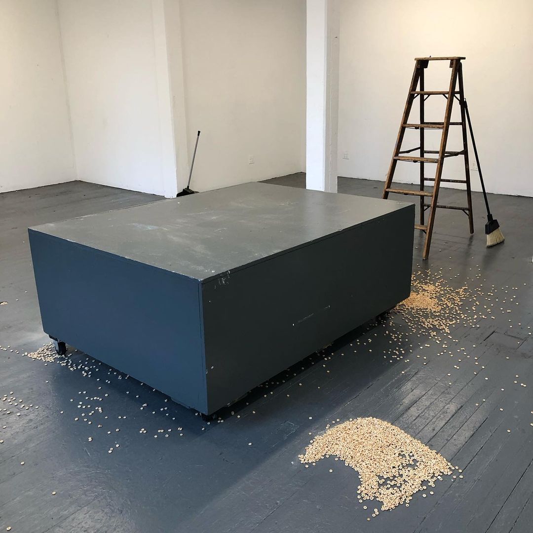 Gray display table with piles of oat-Os cereal on the floor next to it, ladder, and broom