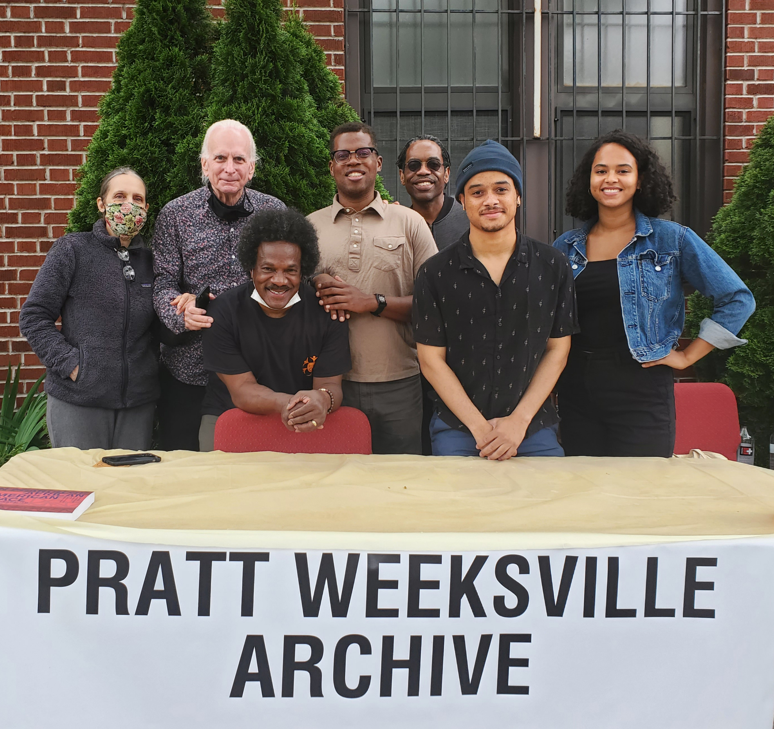 Seven people pose behind an outdoor table reading Pratt Weeksville Archive