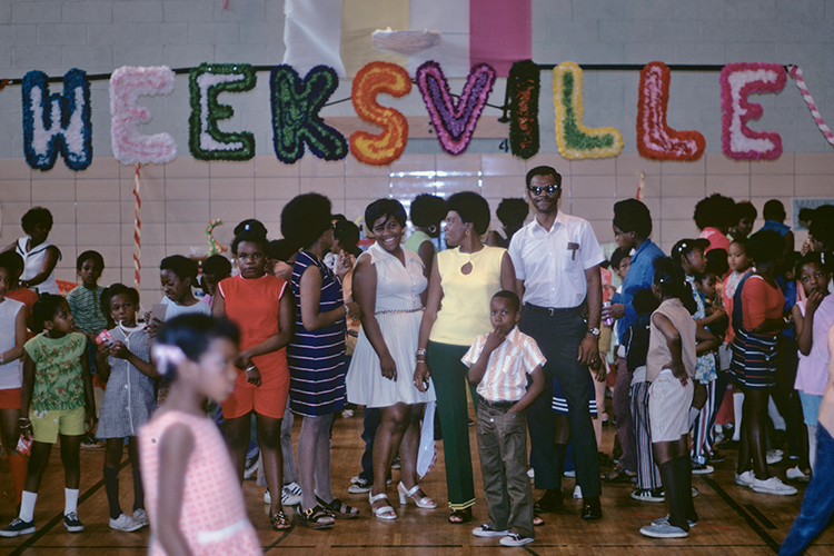 Children and adults gather, smiling, under shiny puffy letters spelling Weeksville
