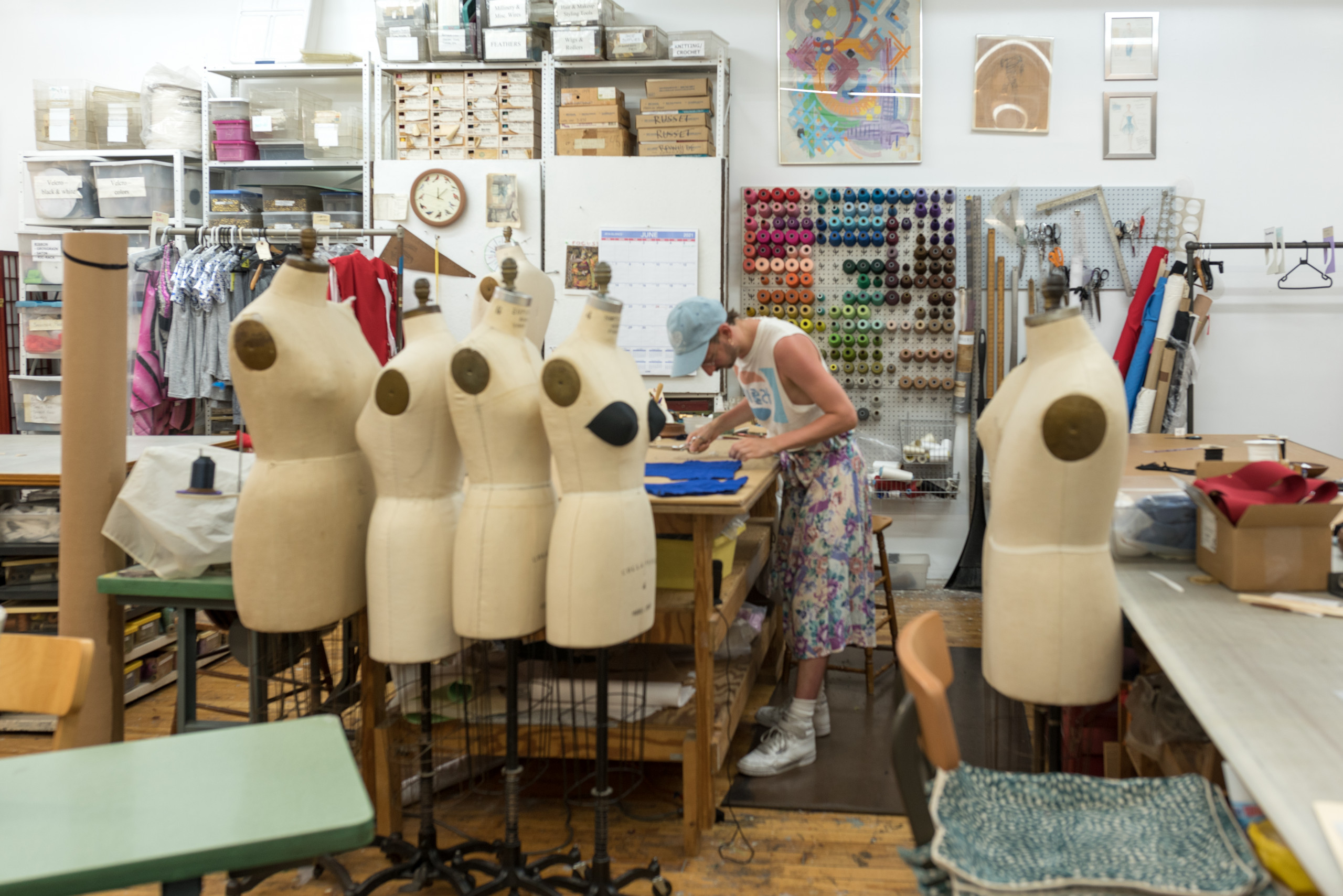 Garment maker works with pattern pieces at table behind row of dress forms