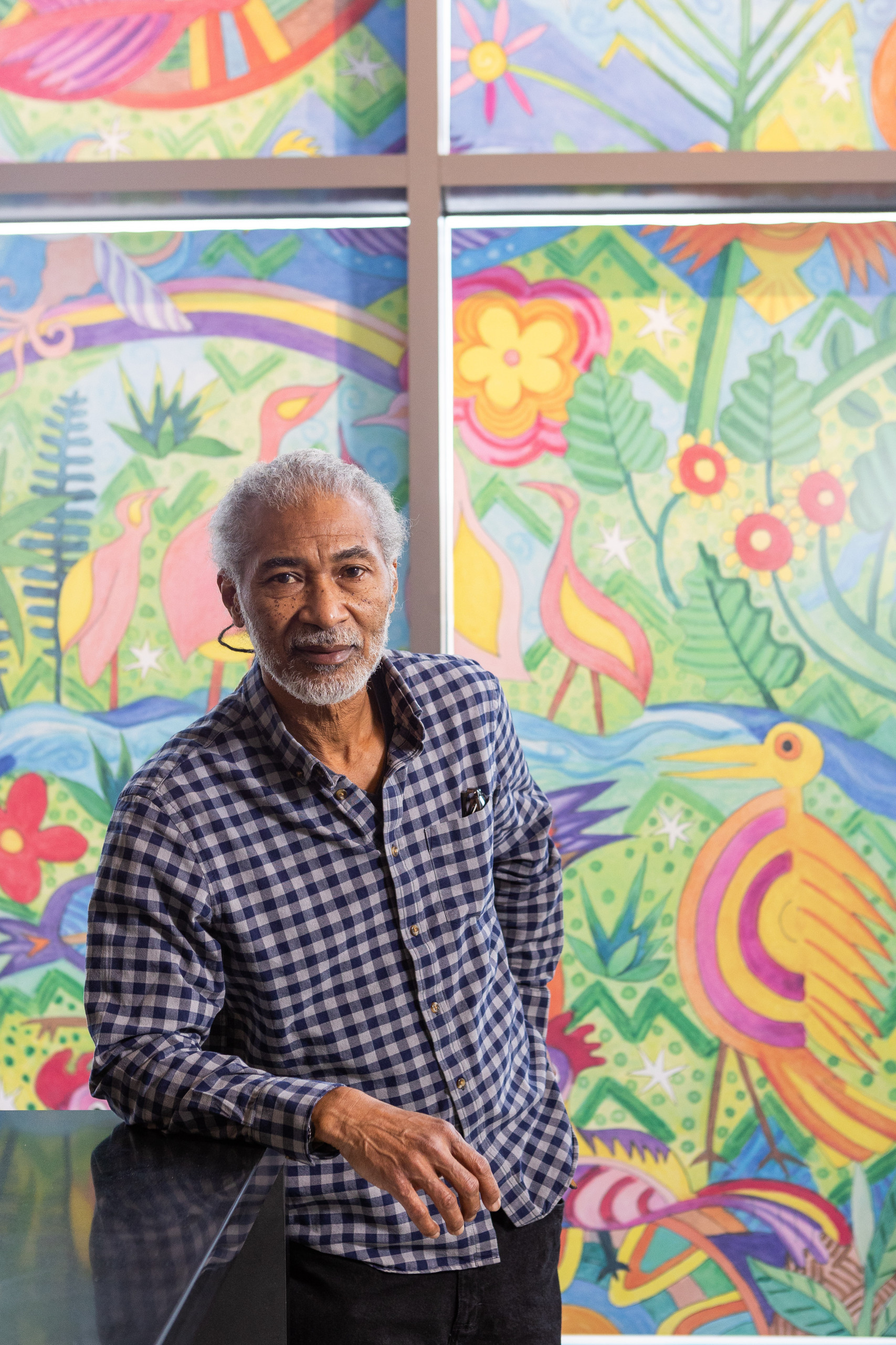 Artist Noël Copeland poses in front of a colorful window painting of vegetation and birds