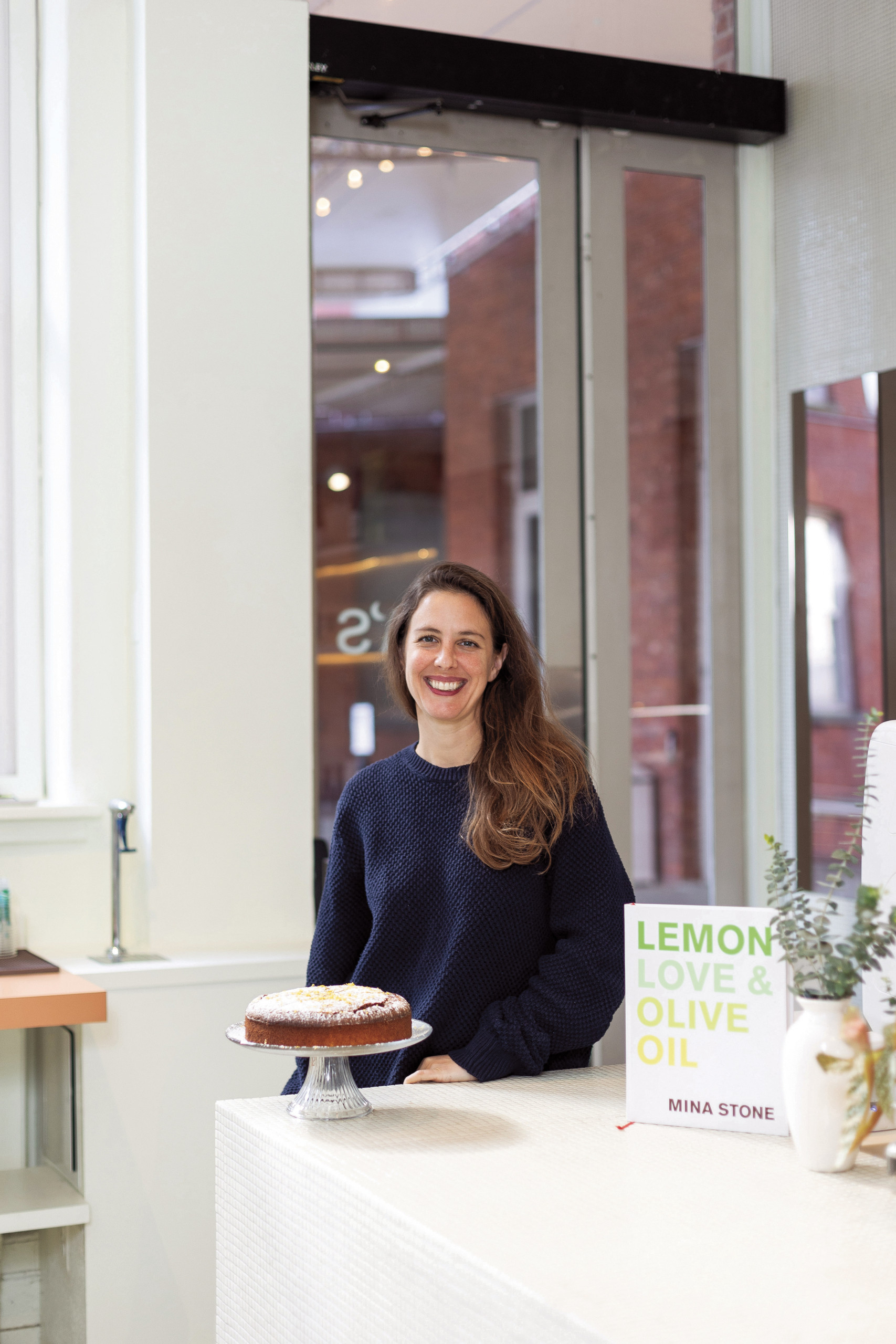 Mina Stone poses at the counter of her café behind a lemon cake and a copy of her book Lemon, Love, and Olive Oil