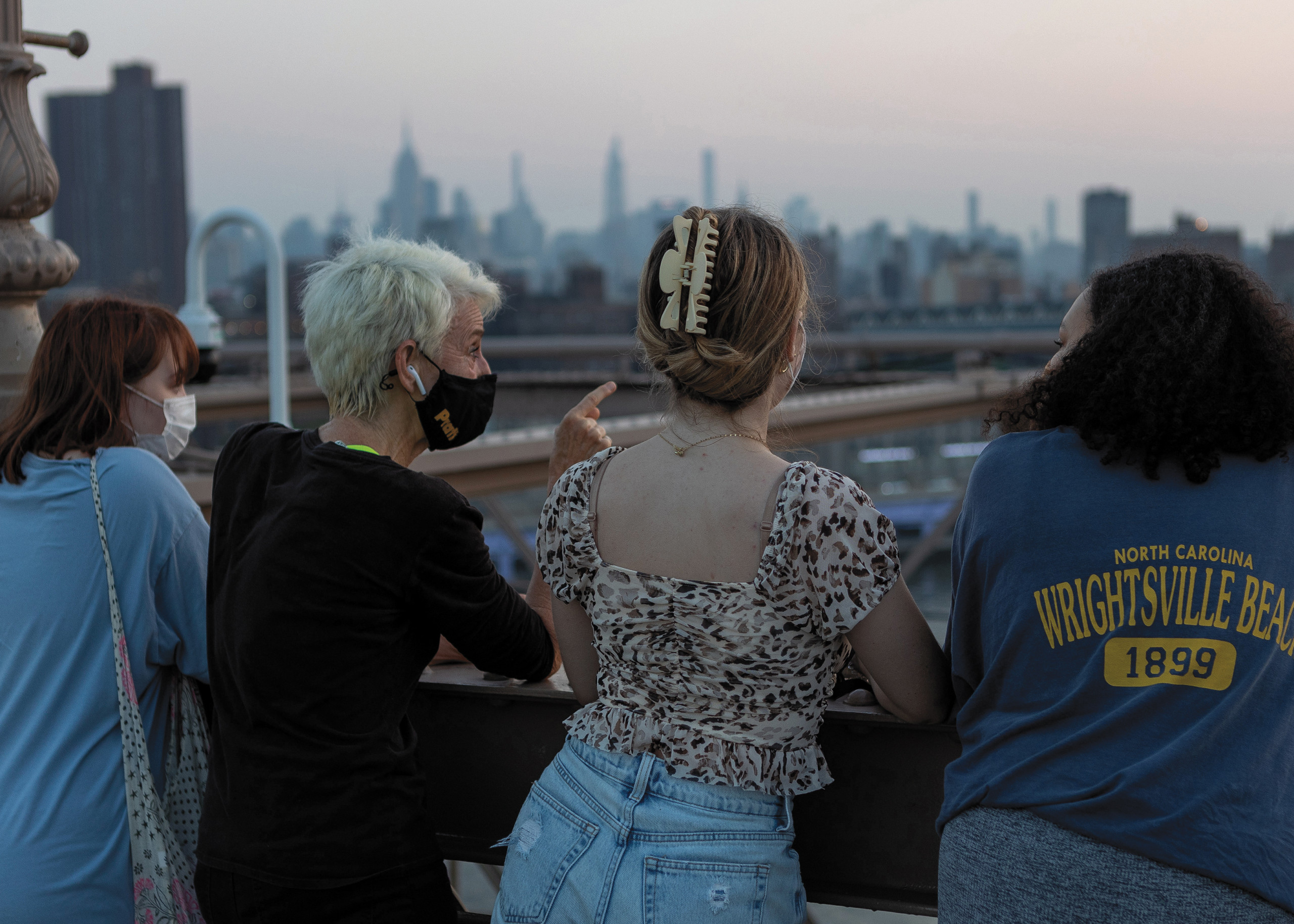 Three students flank President Frances Bronet, who points to an object out of the frame, looking at the city skyline from the Brooklyn Bridge