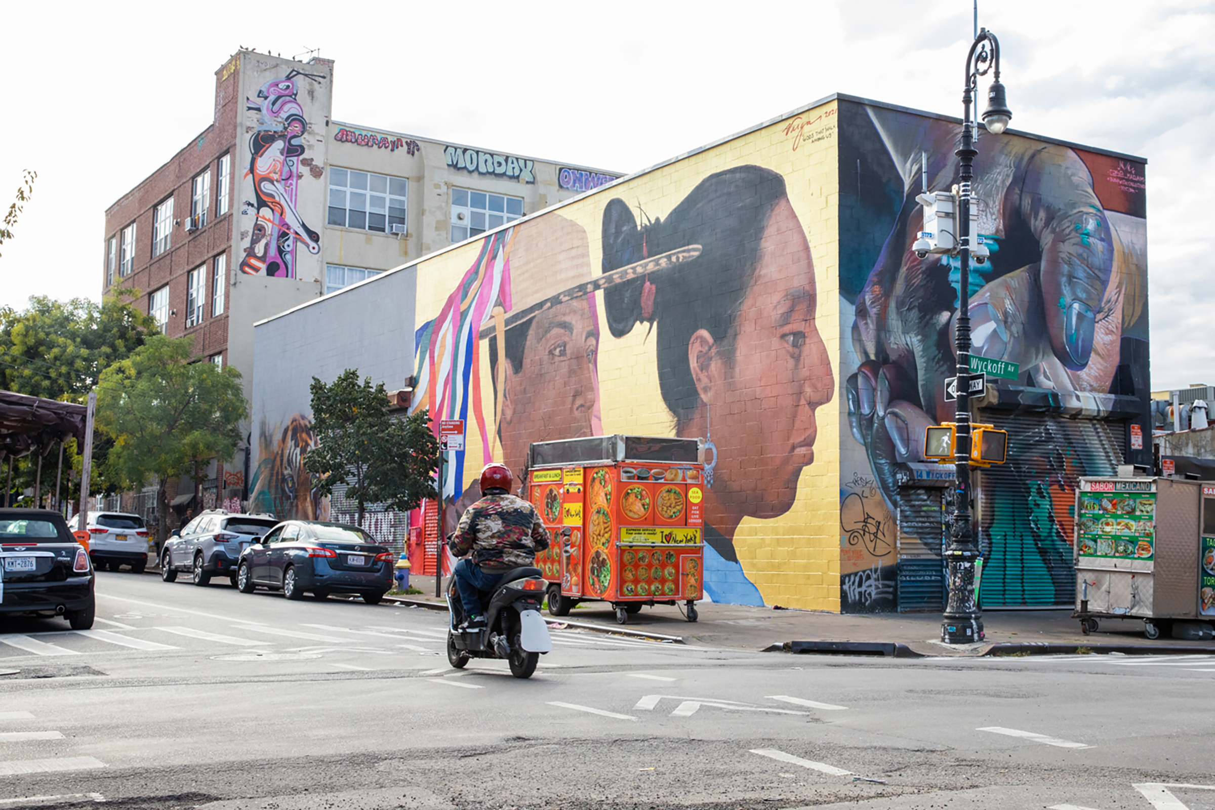 Mural with two brown-skinned people, one wearing a brimmed hat with colorful ribbons, the other with dark hair pulled in a bun and blue circular earring, on a street with other wall art