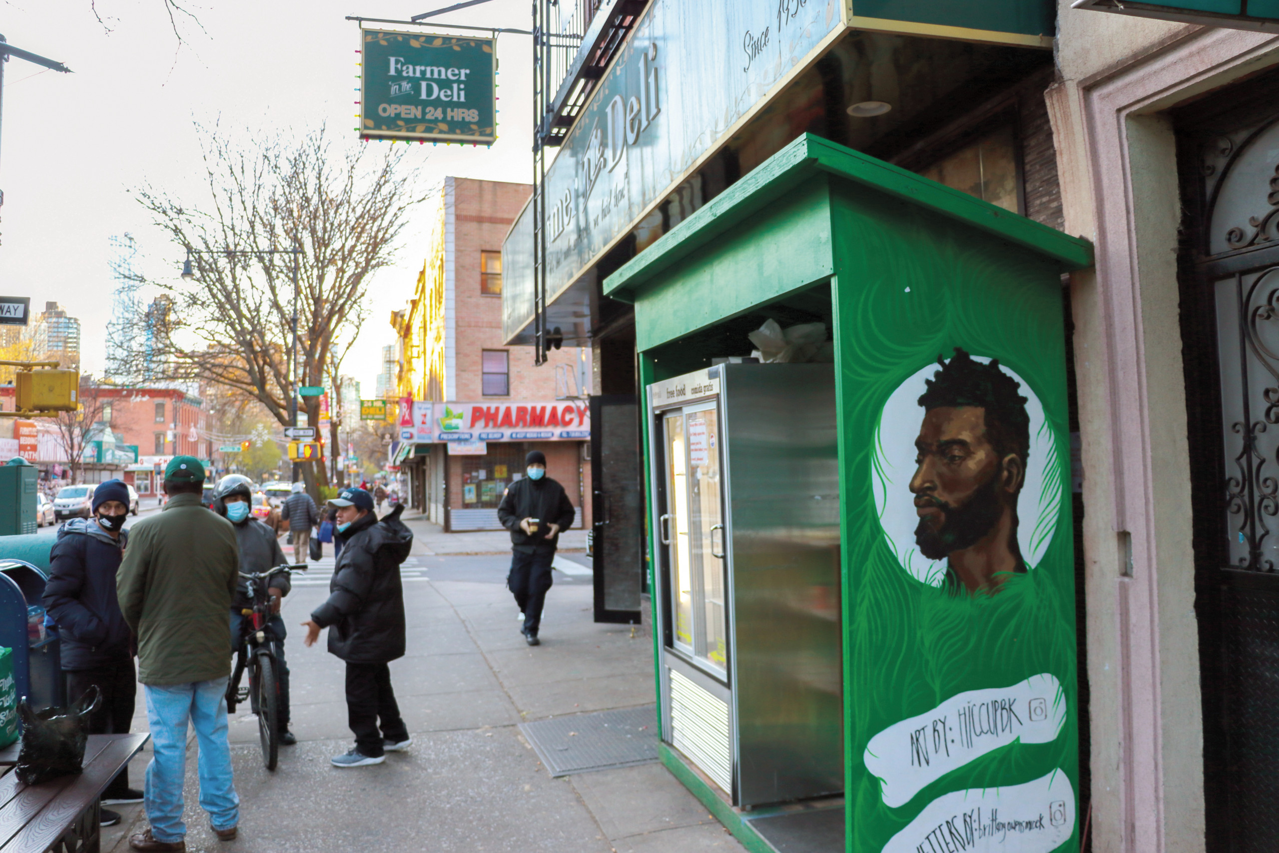 Delivery workers and pedestrians on the Fort Greene street next to a community fridge shelter painted green with a portrait of a brown-skinned person encircled in white