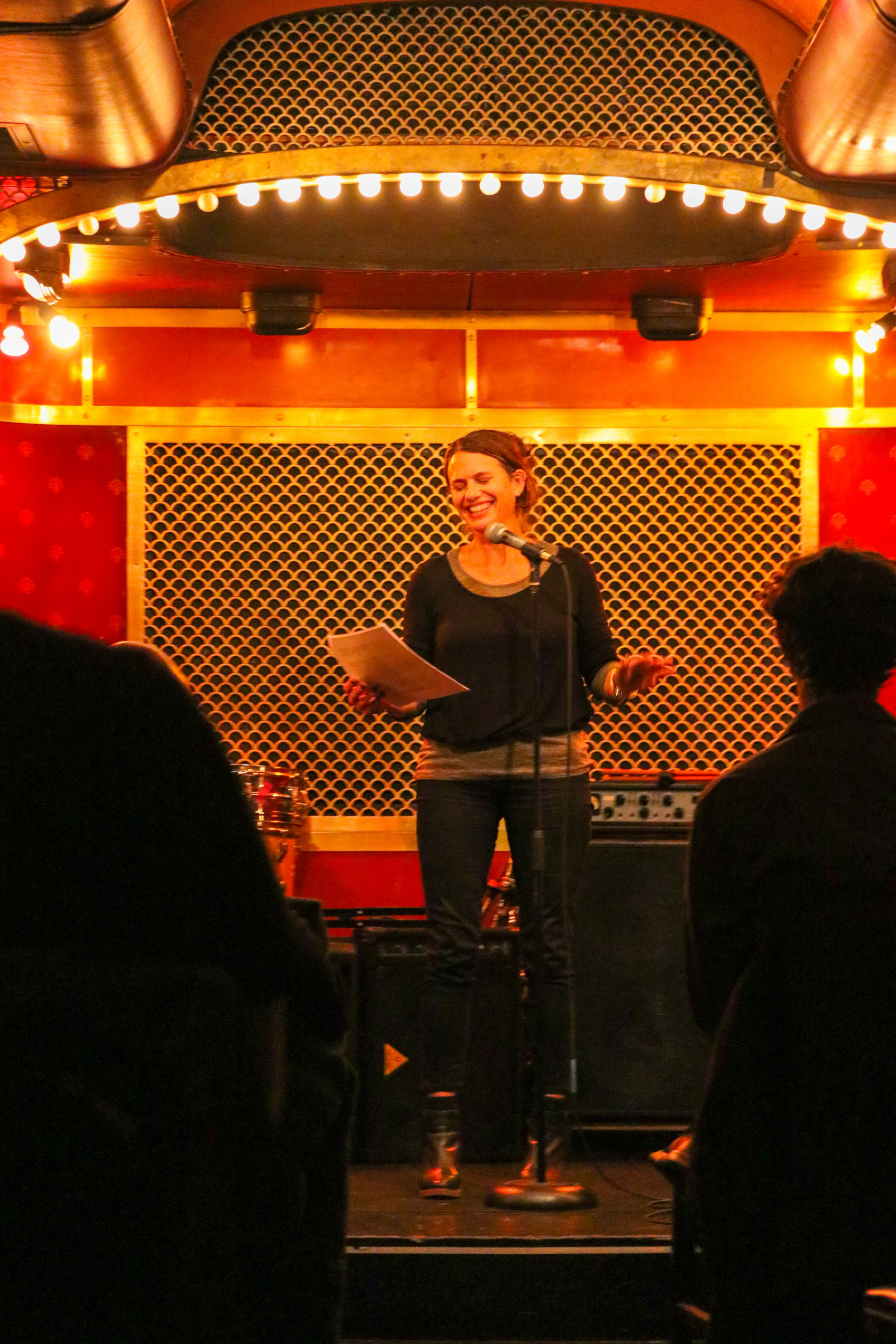 Author Anna Moschovakis, smiling, reads from pages on a stage beneath an arc of marquee lights