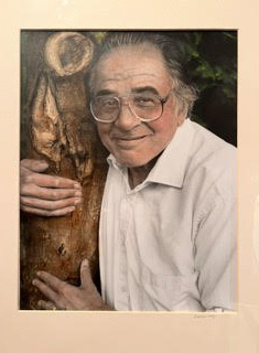 Person with dark and light gray hair, large-framed glasses, a white button-down, and a smiling expression wraps his hands around a knotty tree trunk