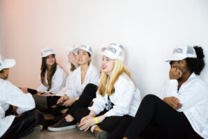 students sitting a room, wearing matching white hats that say "fashion", seated on the floor of a room
