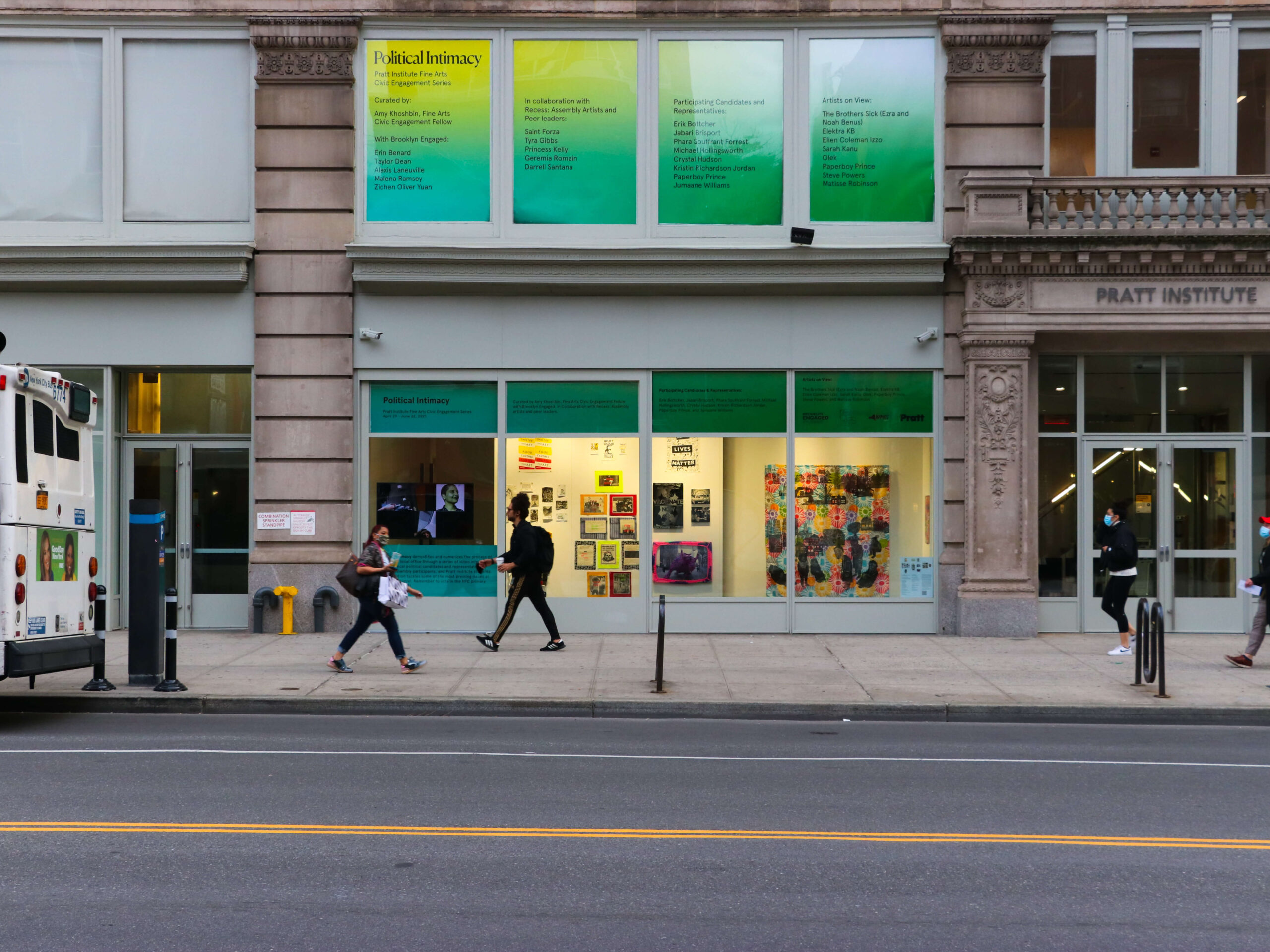 Photo of the façade of a building. It is is green and yellow with large windows. There are people walking on the sidewalk directly in front of it. Part of a bus can be seen at the edge of the image.