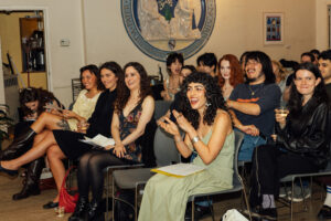 A diverse group of people is sitting in a room, smiling and enjoying themselves. They appear to be attending an event or performance. The foreground features several women, one in a green dress, clapping with enthusiasm. Others show expressions of laughter and engagement. Various drinks are present on the tables, and there are notes and papers in hand. The background includes a decorative wall with a circular emblem.