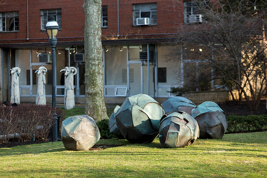 metal sculpture spheres rest in the grass with a tree, a building and a set of goat-like statues in the background