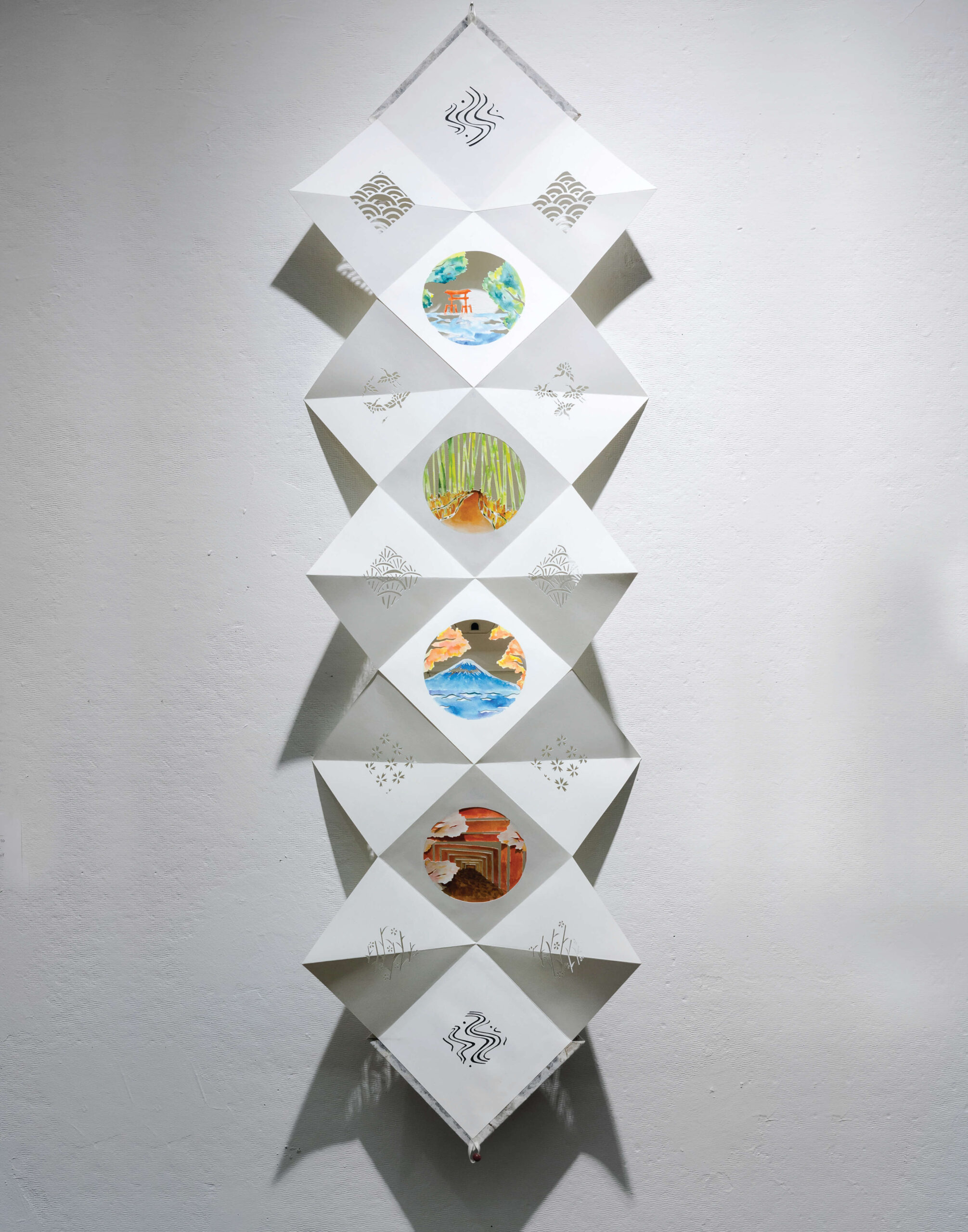 An origami paper sculpture with a colorful drawing along triangular faces is hung against a white wall.