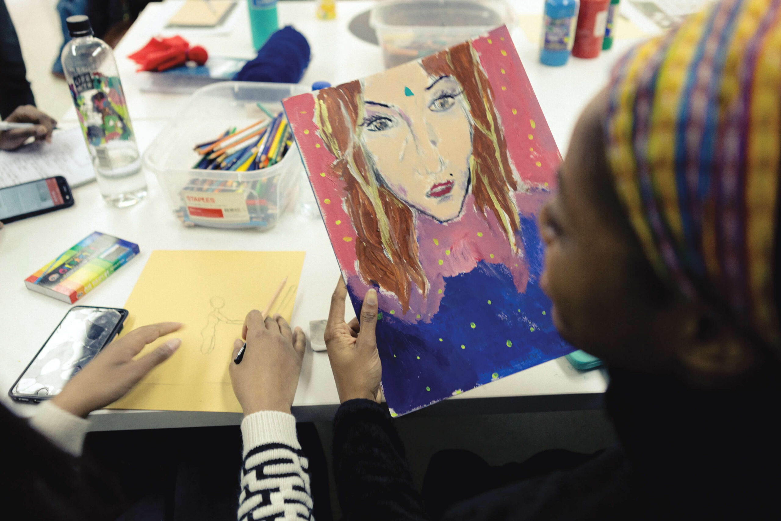 A photo taken behind a student as they hold their art piece while sitting on a desk.