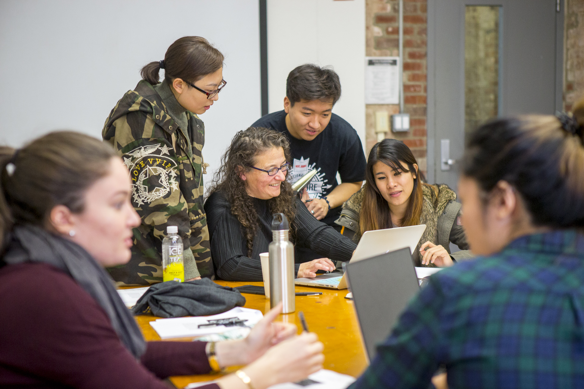 A group of young people are gathered around a teacher as they look at her computer screen. They are in a classroom with a grey door in the background.