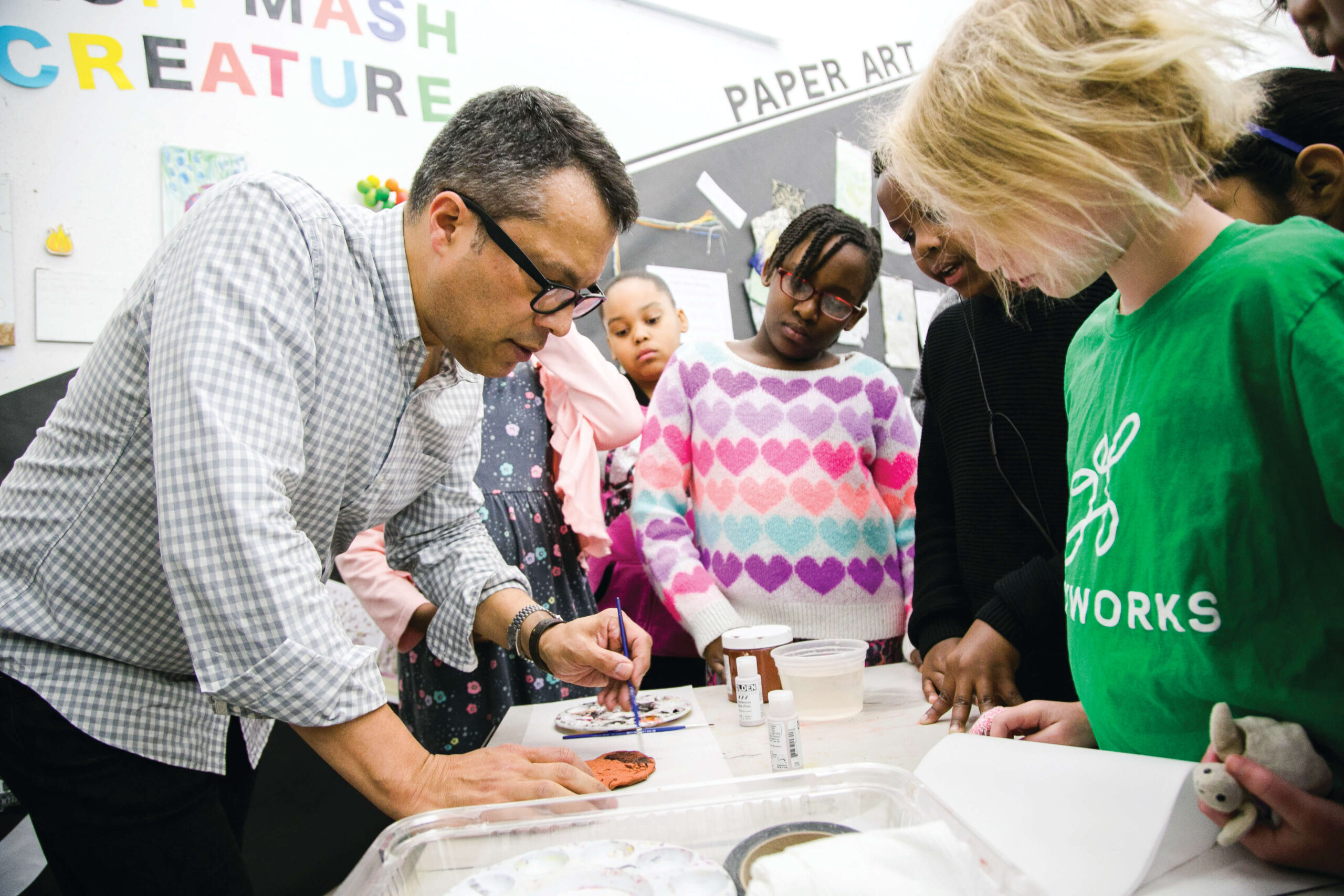 A teacher instructs children around a table. He is drawing with a pen on the table.