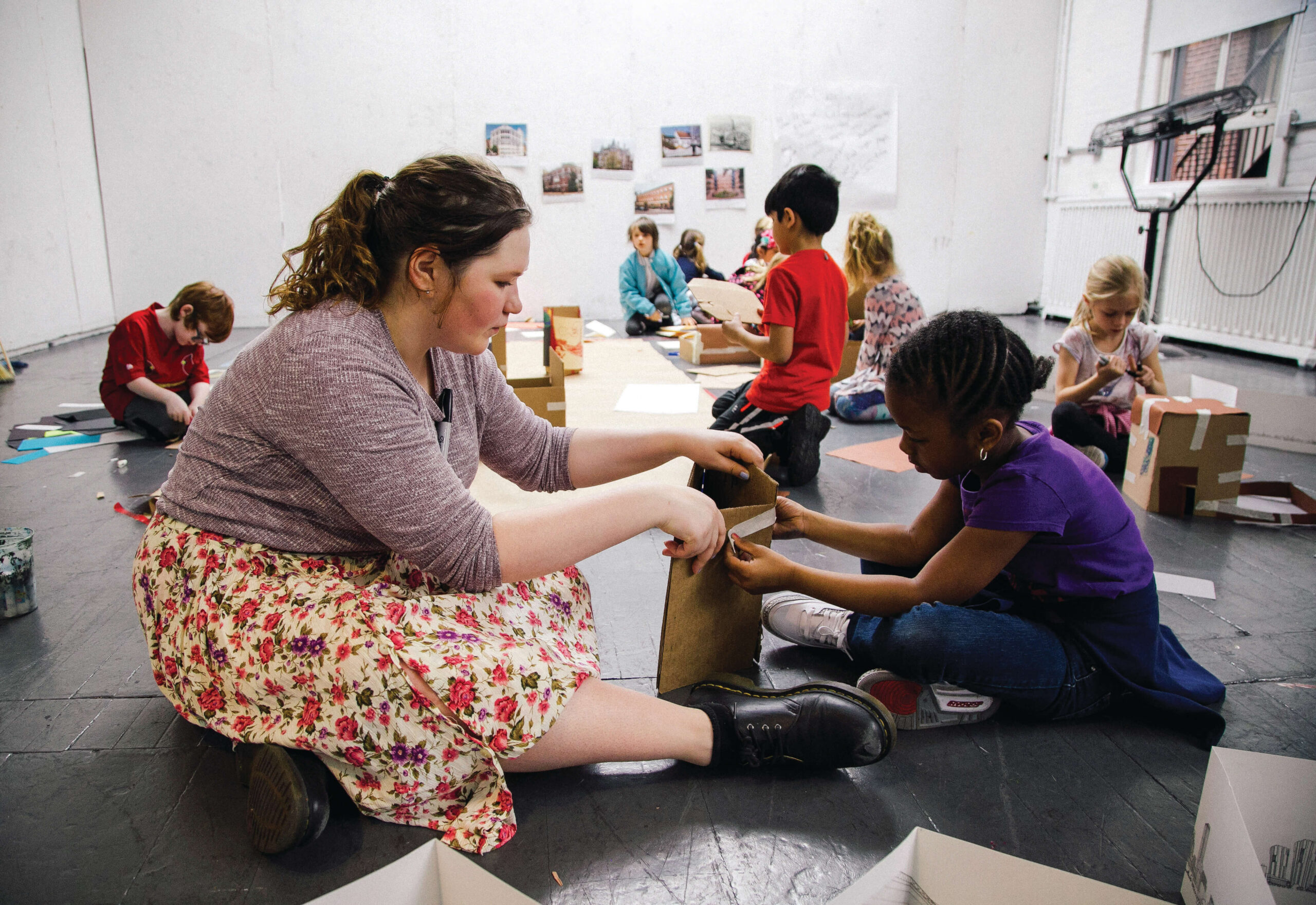 A graduate student sits on the floor to assist a child with their cardboard art project.