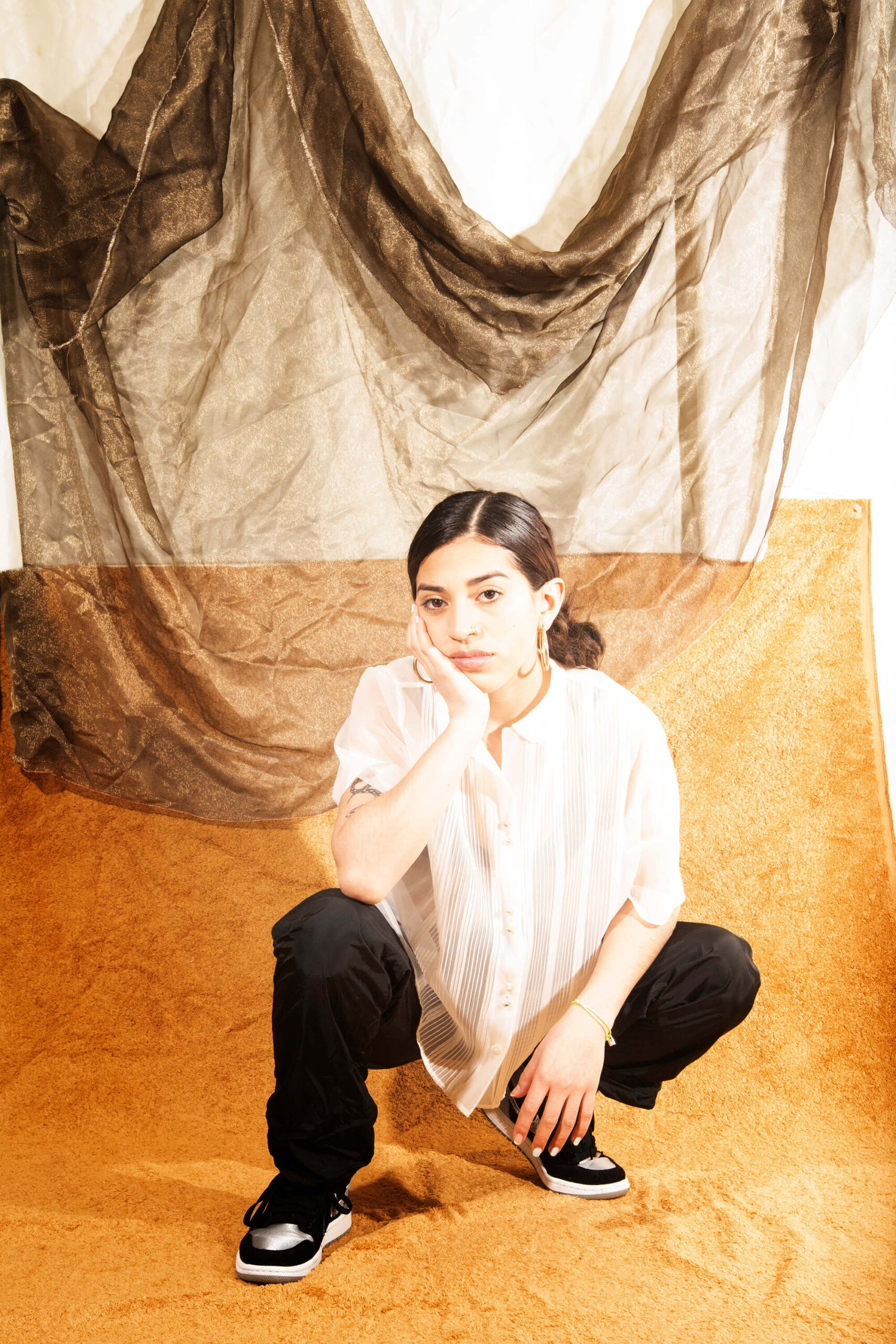A subject squats while posing for a photo on wooden floor. There is a large cloth hanging from a white wall behind her.