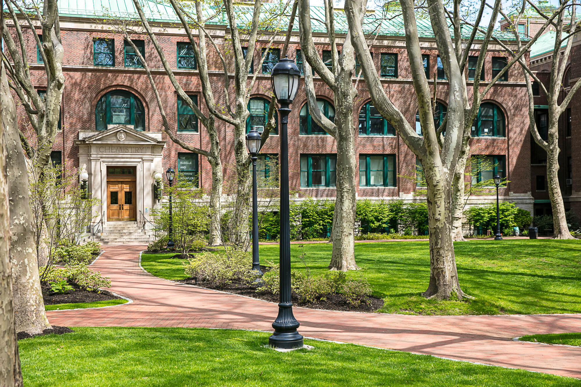 Photo of an empty brick path with a Victorian street lamp and trees surrounding it. There is a red brick building in the background.