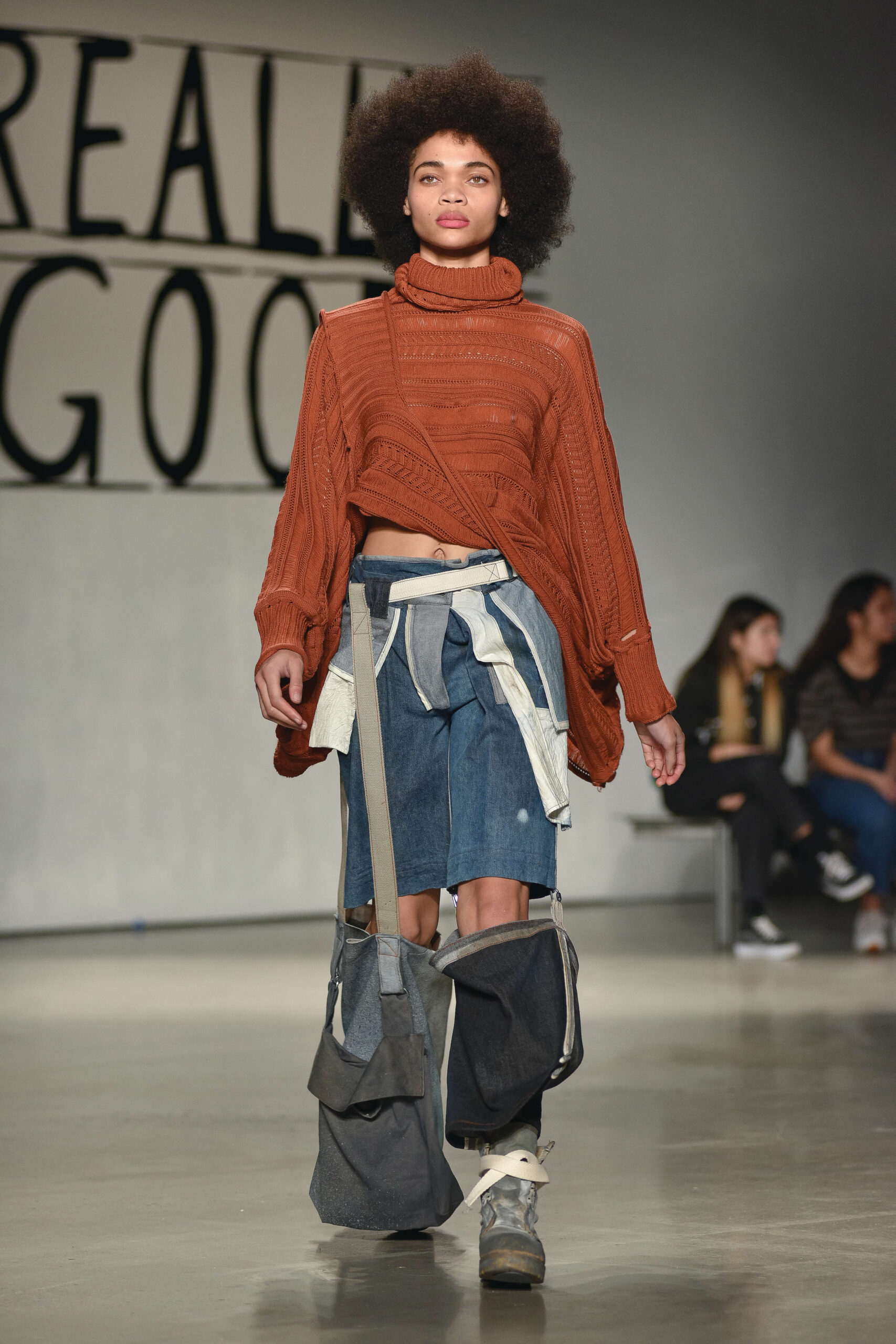 A model walks down a runway wearing deconstructed jeans and a long, draping sweater revealing part of her waist.