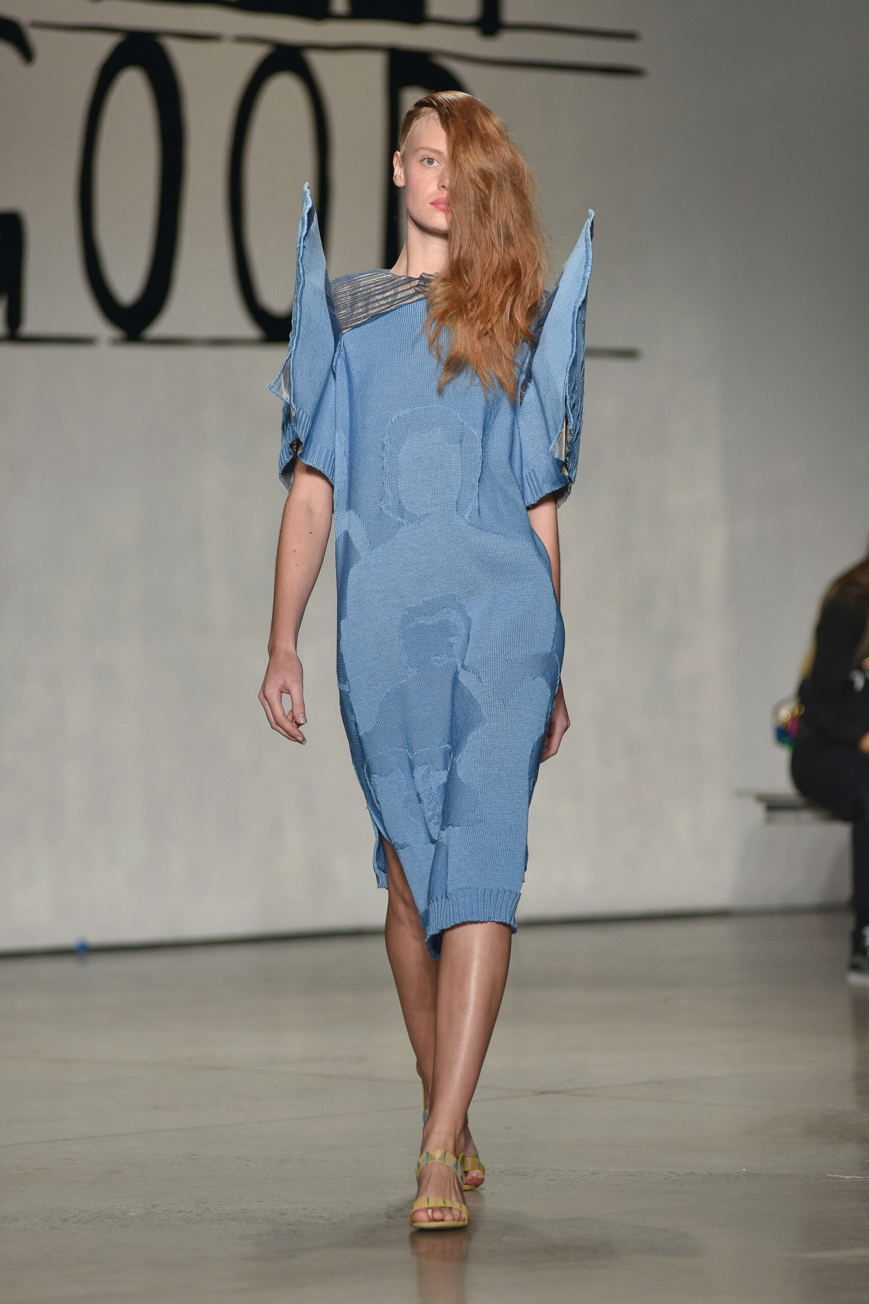 A model walks down a runway wearing a blue dress with should pads that spike upwards.