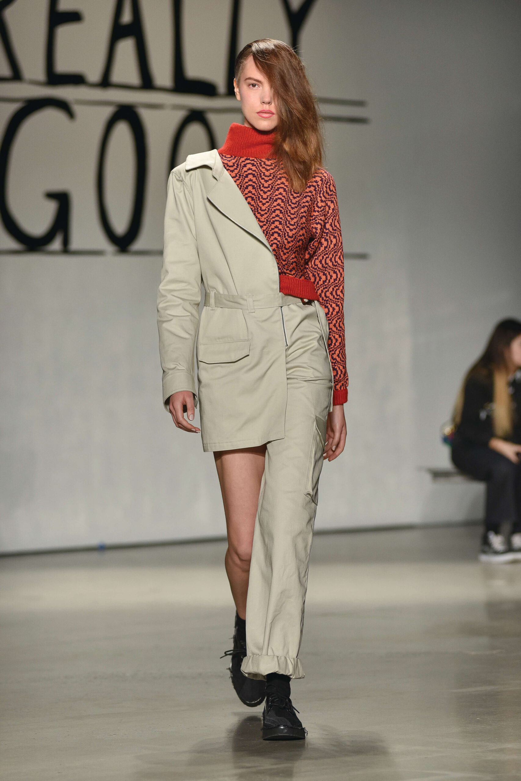 A model walks along a catwalk wearing a beige suit. The pant's right leg is cropped to the length of a short, while the left leg has a normal pant length. The suit's blazer is diagonally divided in beige and red.