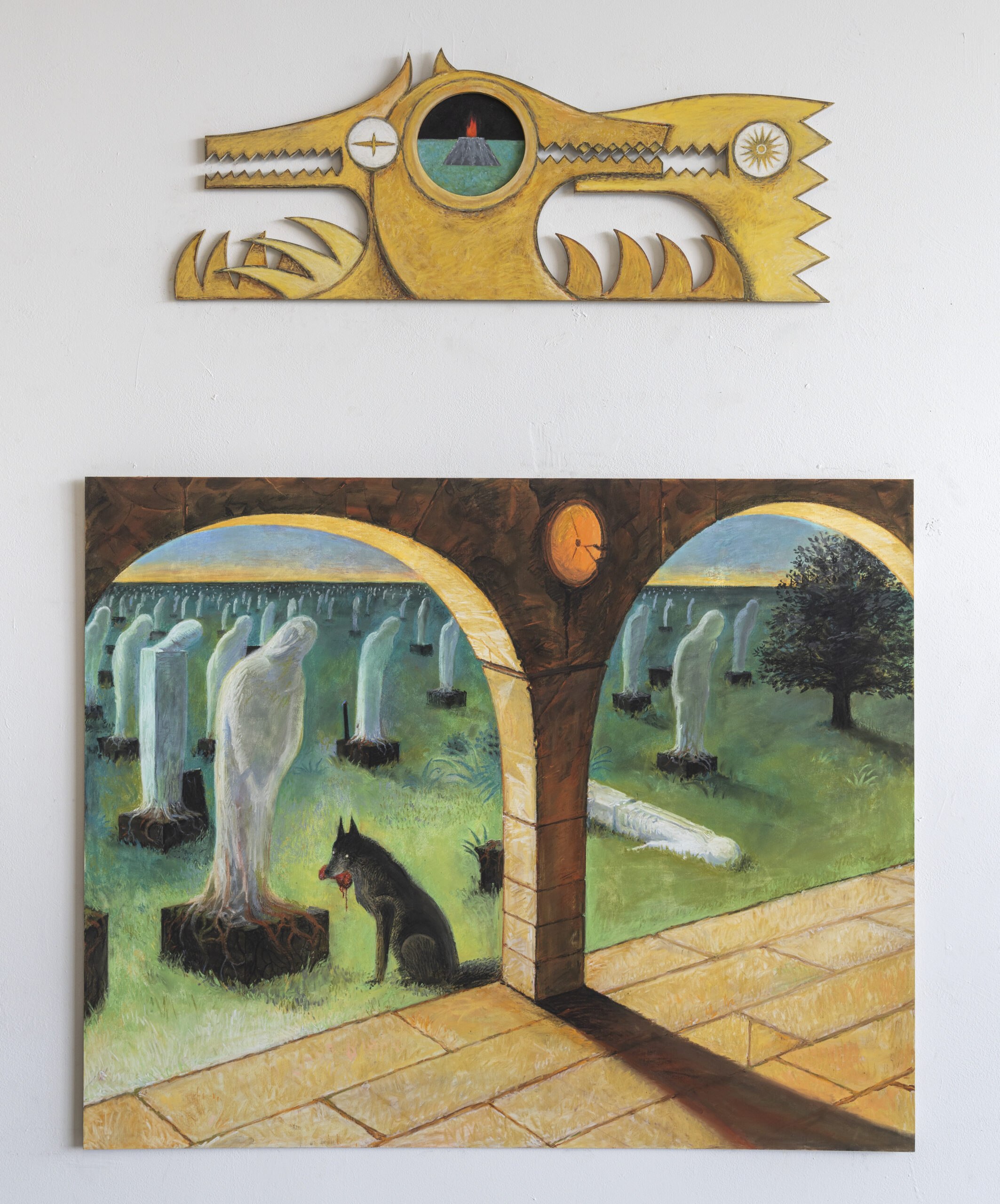 A mixed-media artwork displayed on a white wall. The top piece is a wooden sculpture shaped like a stylized creature, resembling a dragon or mythical beast, with intricate geometric patterns and a central circular motif featuring a small landscape with a volcano. Below the sculpture is a painting depicting an eerie cemetery scene viewed through the arches of a stone structure. The cemetery is filled with ghostly, shrouded figures and tombstones. In the foreground, under the archway, a black dog with red eyes is seen holding a piece of meat in its mouth, adding to the ominous atmosphere of the scene.