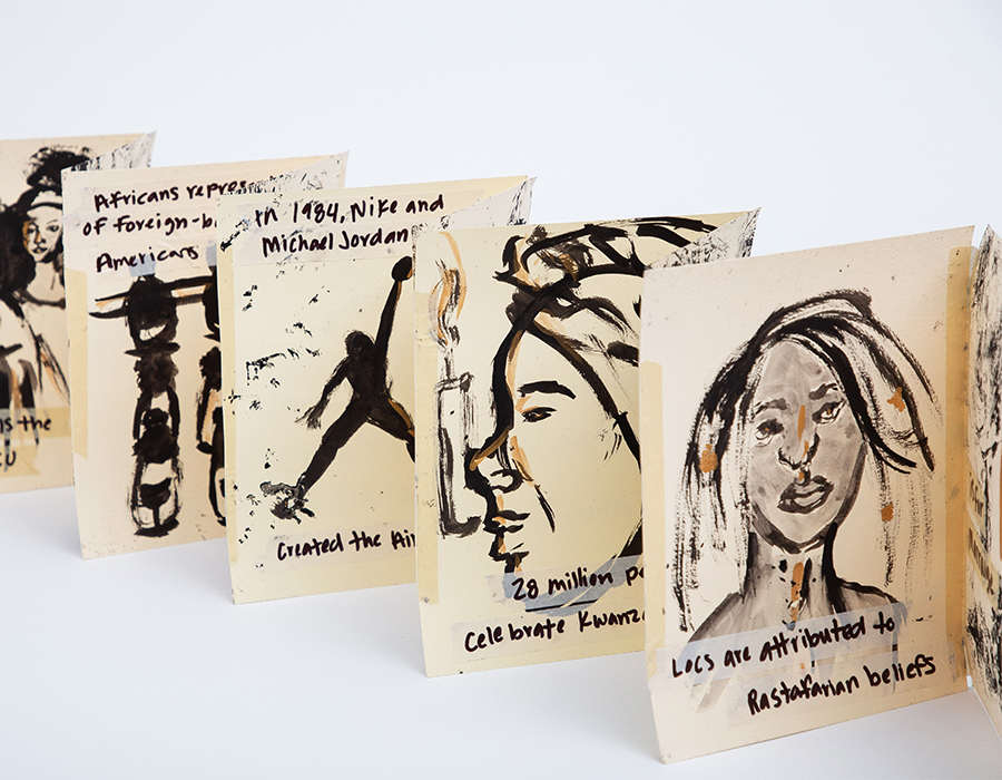 An installation atop a white surface. There are paper pages standing upright with sketches of various black people along writing discussing the black identity,