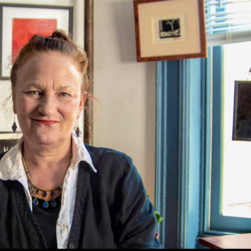 Amy Guggenheim looks directly at the camera with a slight smile. She is standing in front of a white wall with painting hanging from it. There is a window visible to her right