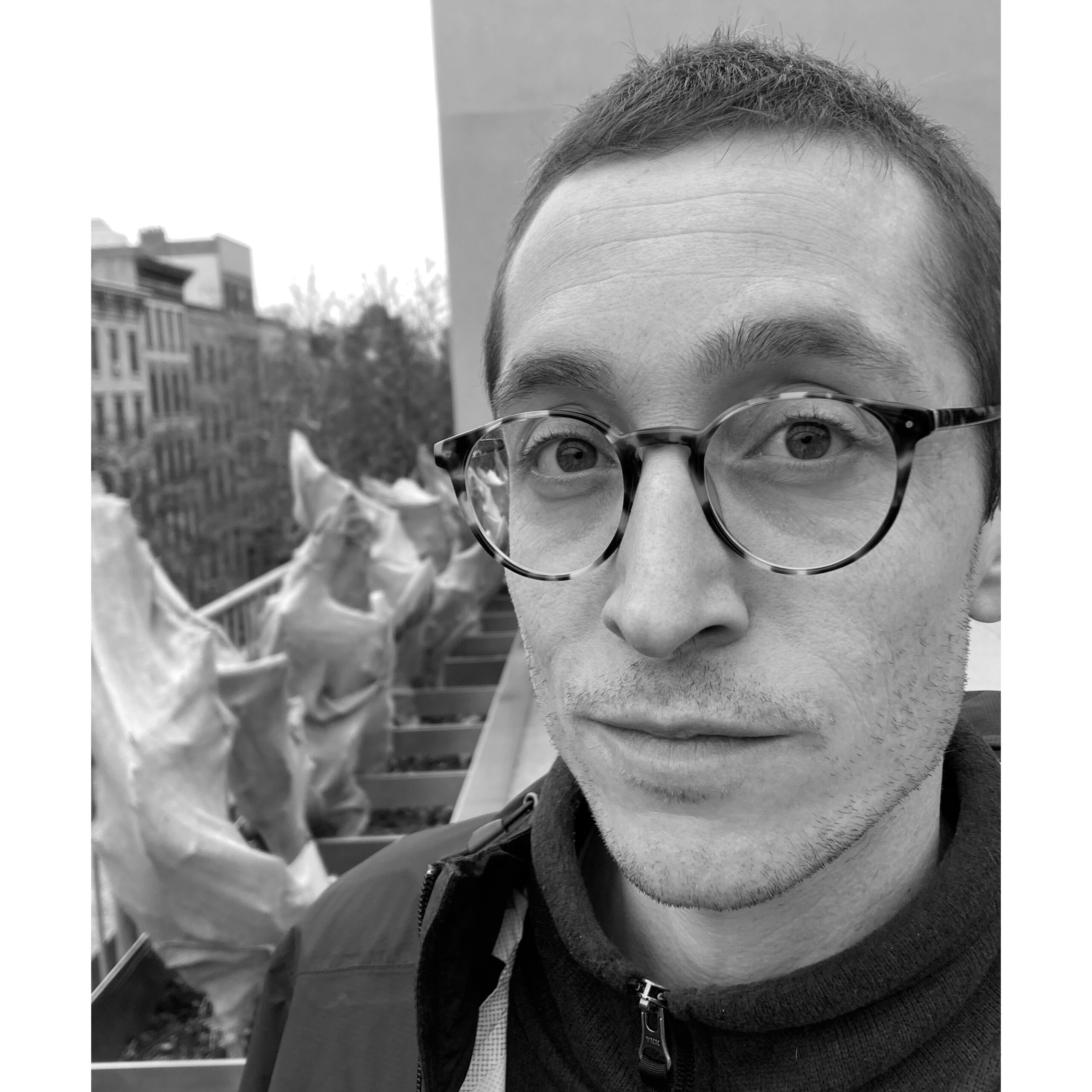 A man with short hair and tortoiseshell glasses stands on a rooftop with sculptural, wrapped plant forms behind him. He wears a zippered jacket and looks directly at the camera with a neutral expression. In the background, New York City buildings are visible. The image is in black and white.