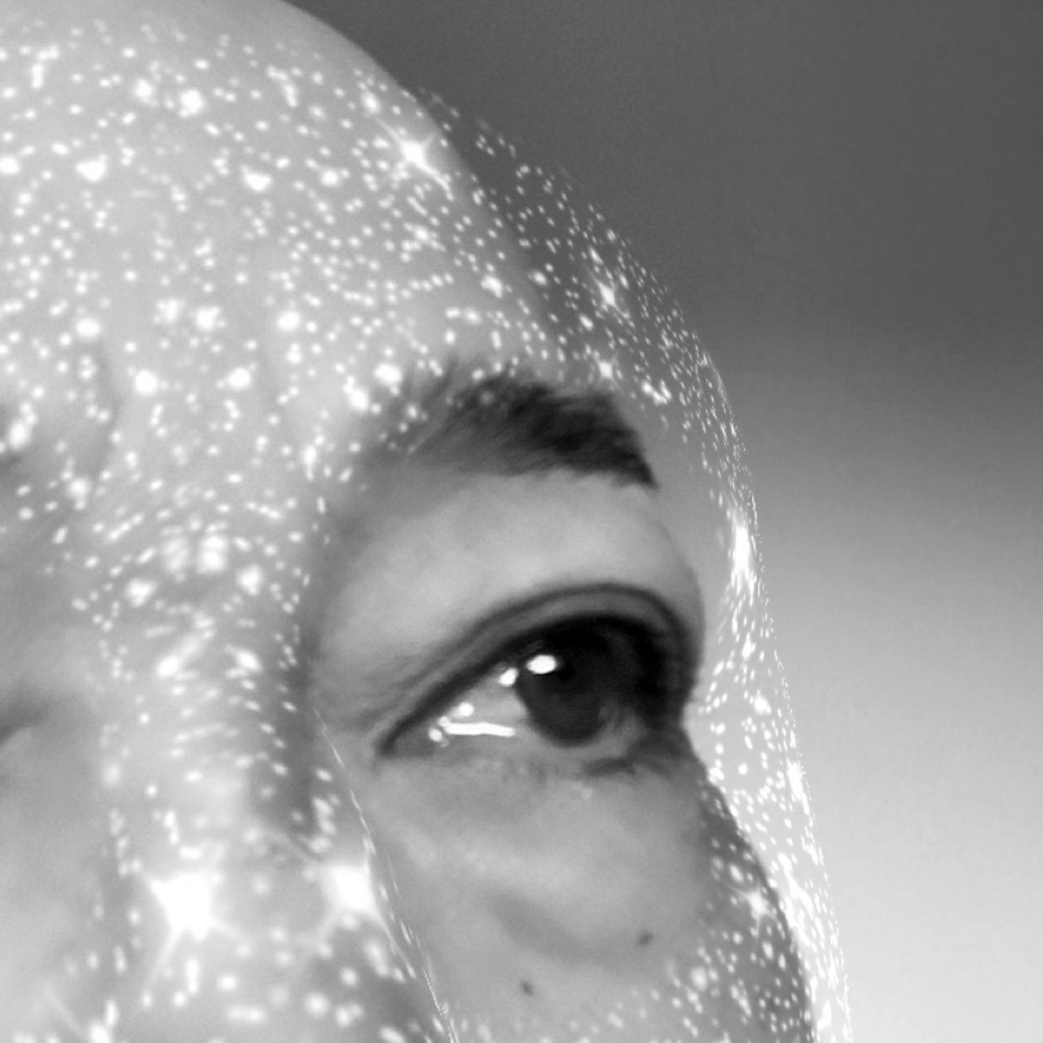 A close-up black and white image of a person’s eye and forehead, with digital starry light projections creating an ethereal and futuristic overlay across their bald head and skin. The image evokes a sense of wonder and introspection.