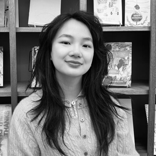 A young woman stands in front of a bookshelf, smiling gently. She wears a cable-knit cardigan and has long, layered hair. The shelves behind her display illustrated books, adding a literary and artistic context.