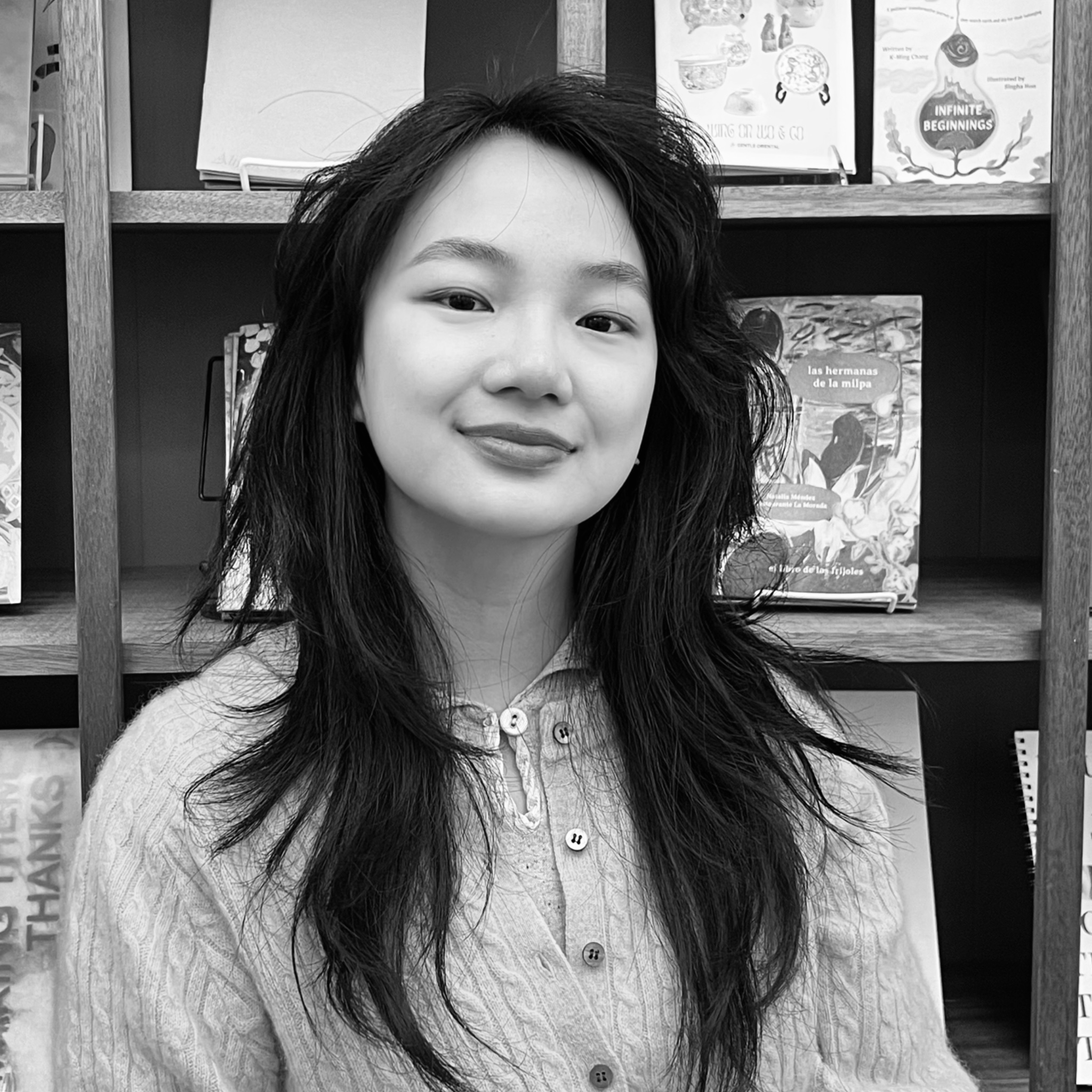 A young woman stands in front of a bookshelf, smiling gently. She wears a cable-knit cardigan and has long, layered hair. The shelves behind her display illustrated books, adding a literary and artistic context.