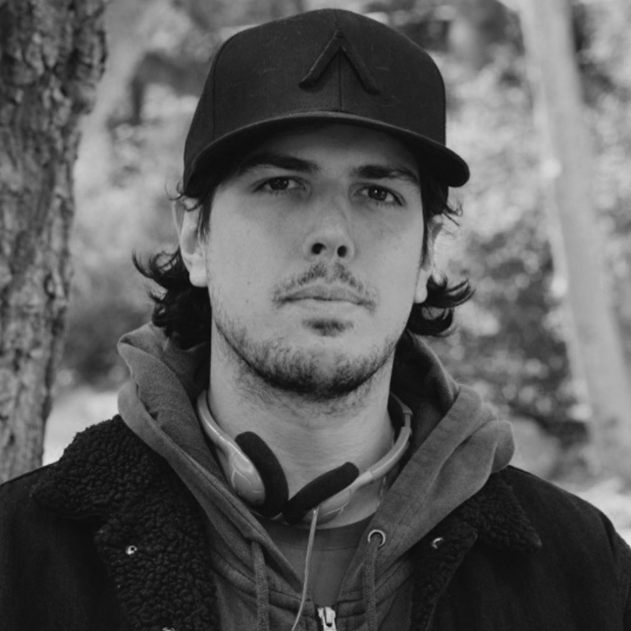 A man stands outdoors wearing a dark cap, hoodie, and jacket with headphones around his neck. His expression is serious, and he is positioned next to a tree, with a blurred forest background. The image is black and white.