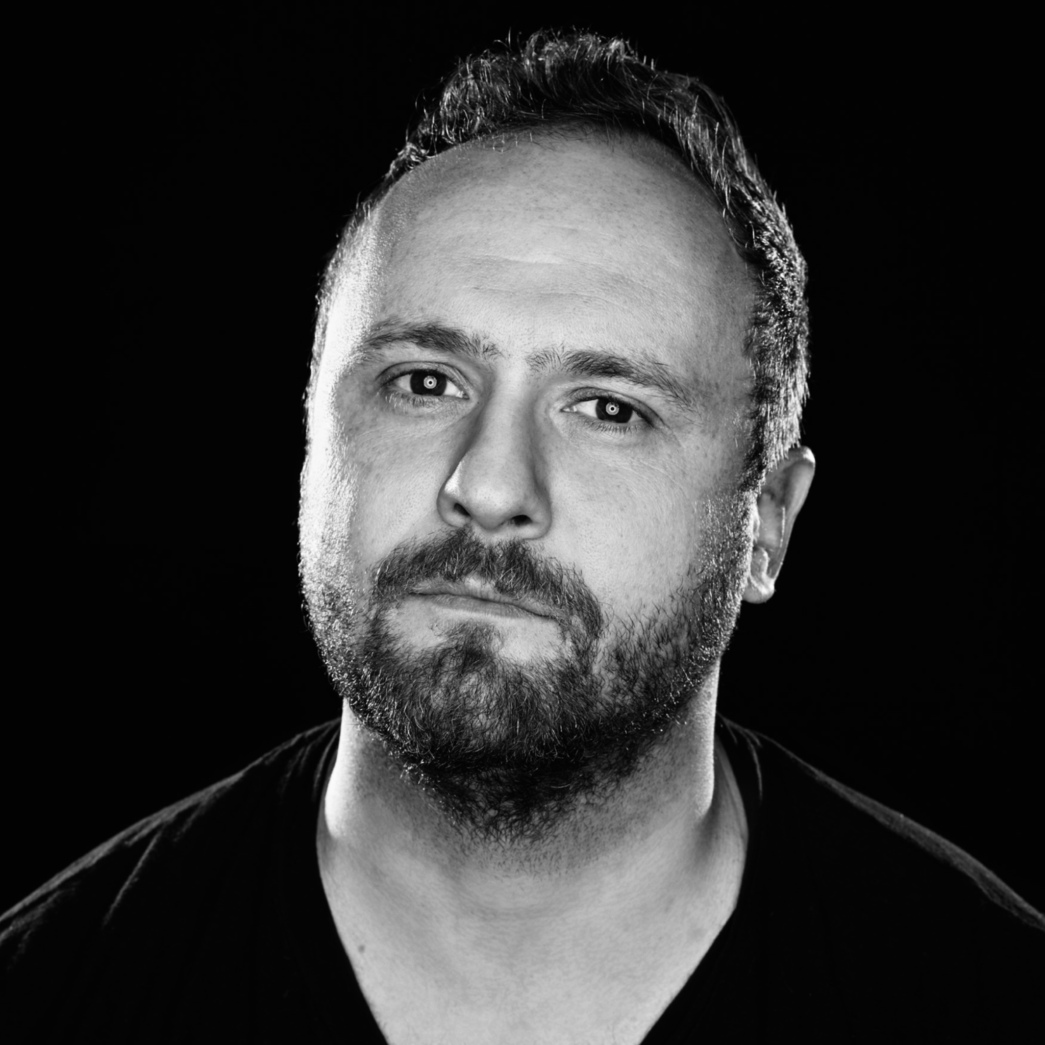 A man with a short haircut and a trimmed beard gazes directly at the camera with a neutral expression. He wears a dark V-neck shirt and is lit dramatically against a solid black background. The portrait is in black and white, emphasizing contrast and mood.