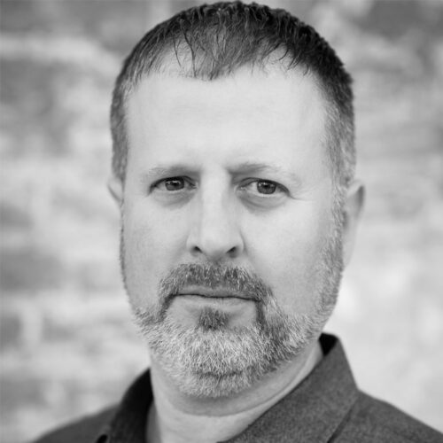 A man with short-cropped hair and a salt-and-pepper beard stares intently at the camera. He wears a dark button-up shirt, and behind him is a textured stone wall. The portrait is in black and white, highlighting strong facial features and lighting contrast.