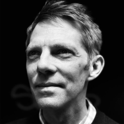 A man with short, light hair gazes slightly off-camera with a thoughtful expression. He wears a collared shirt and sweater. The black background and sharp lighting highlight the contours of his face in this high-contrast black and white image.