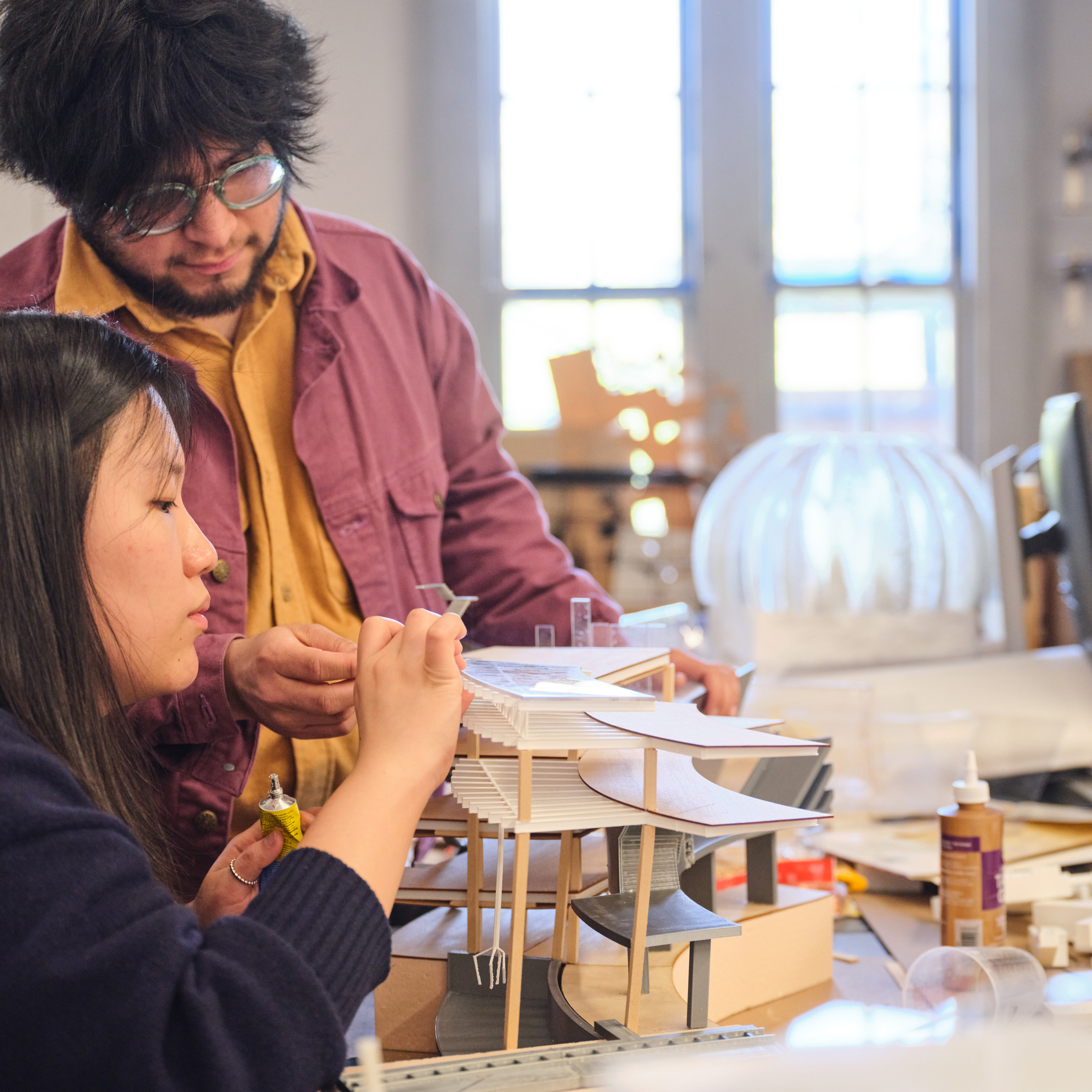 two people discussing what they are working on, in a large studio room, filled with architectural models