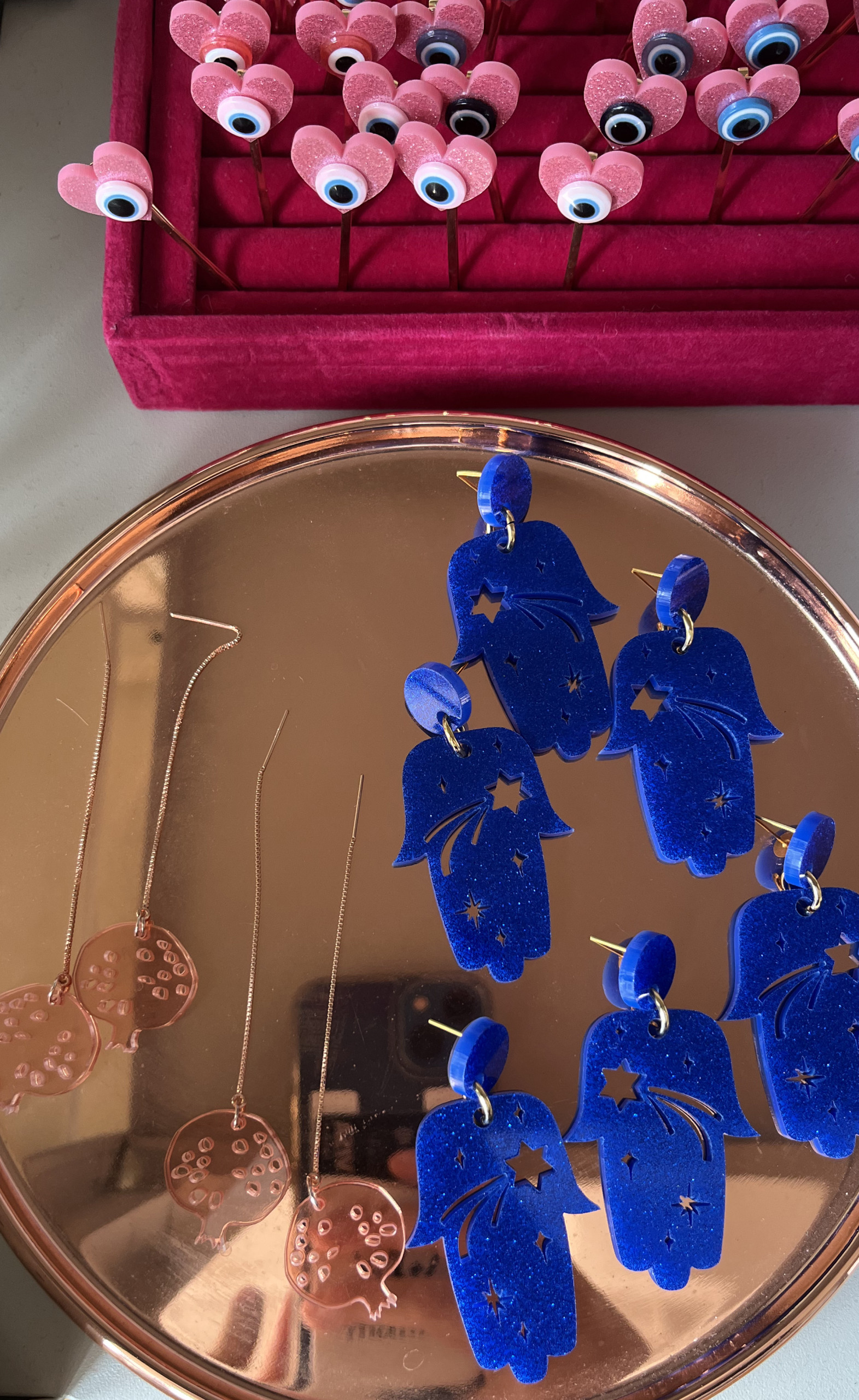 Jewelry pieces on display, pink hearts with eyes, blue hamsas with shooting stars of David, pink pomegranates