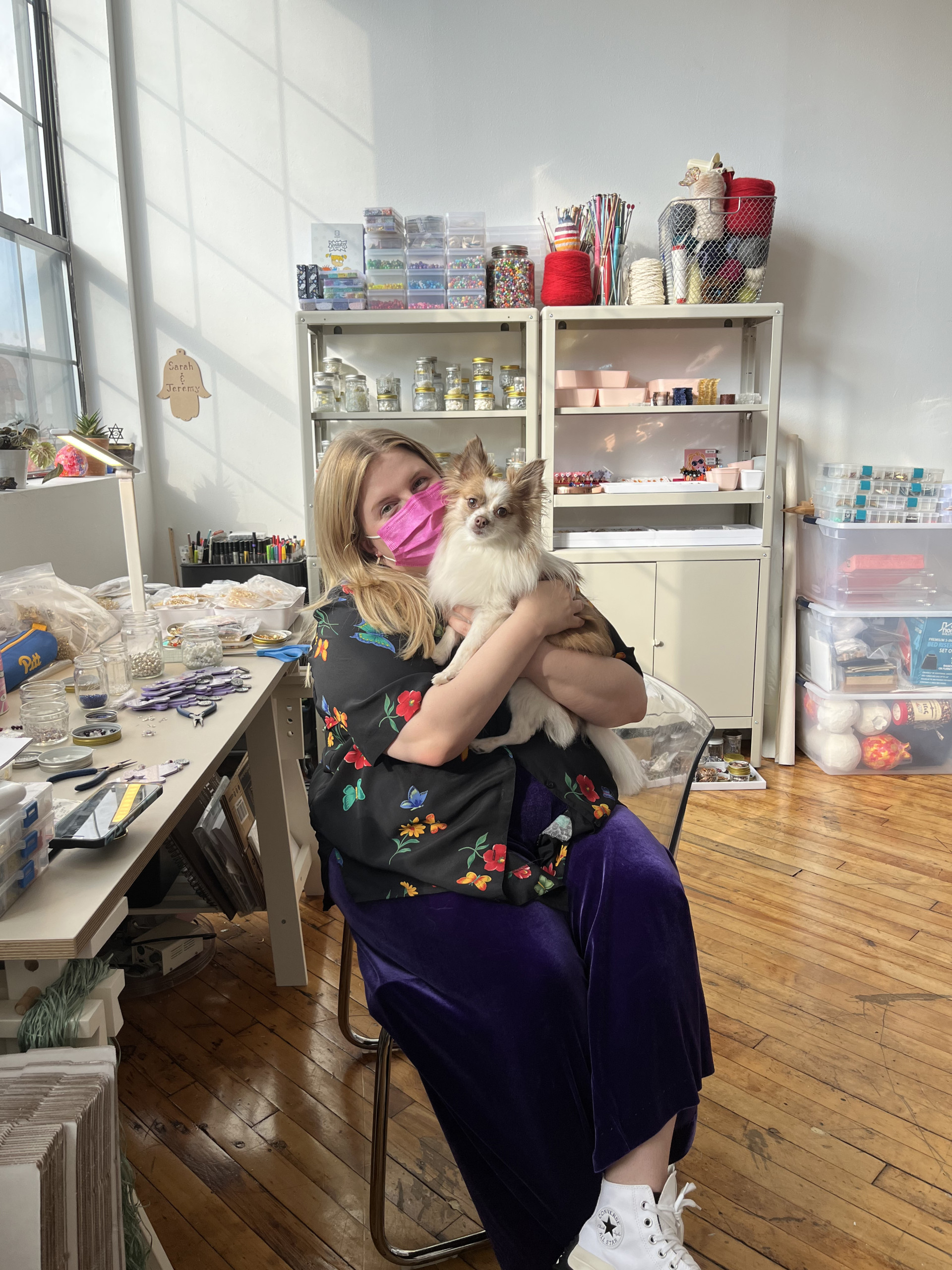 Person with long blond hair, colorful shirt and skirt, and pink mask sitting at work table holding small white and honey-colored dog