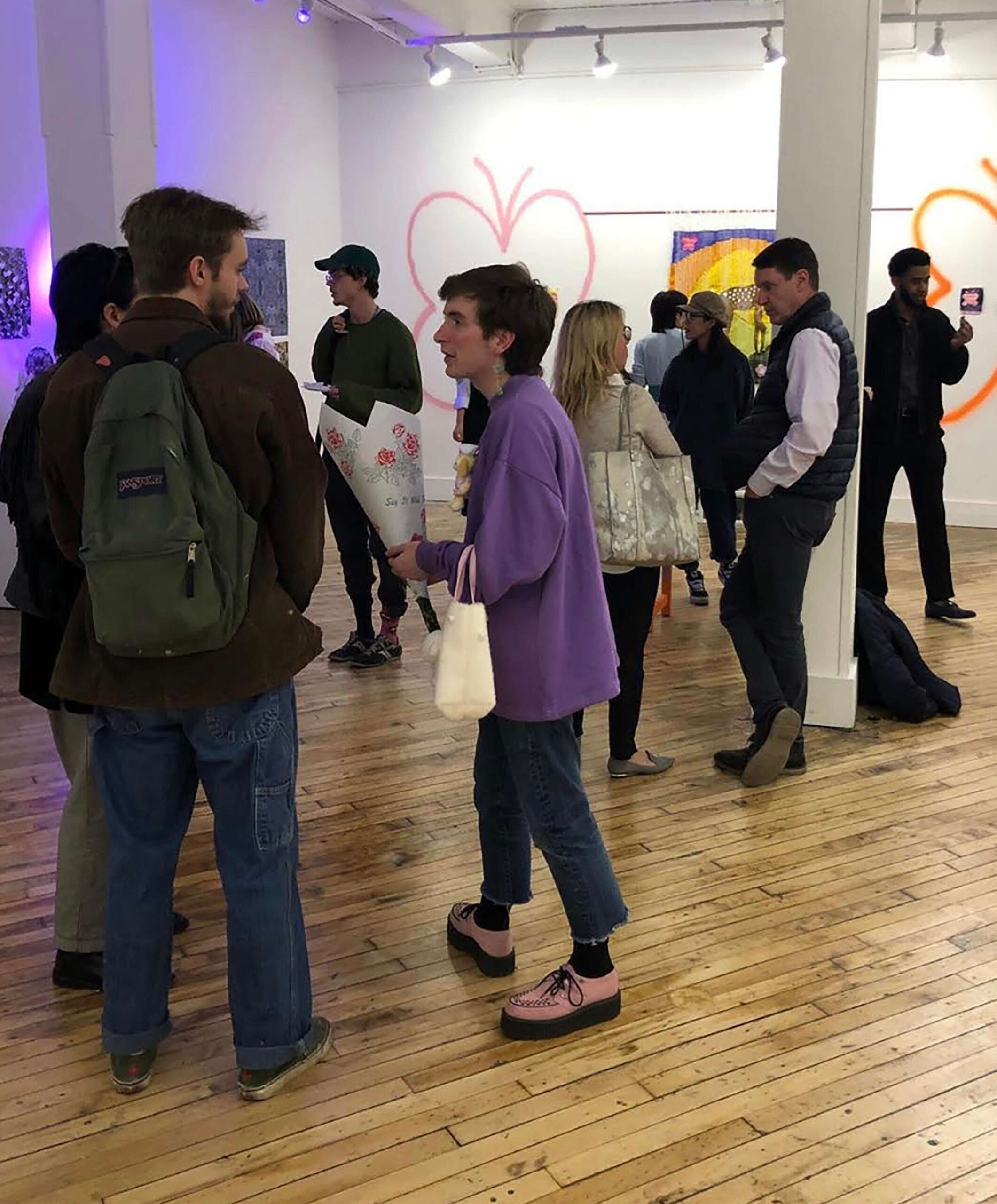 A large group of people chat amidst a large gallery with white walls, wooden floors and art displayed throughout.
